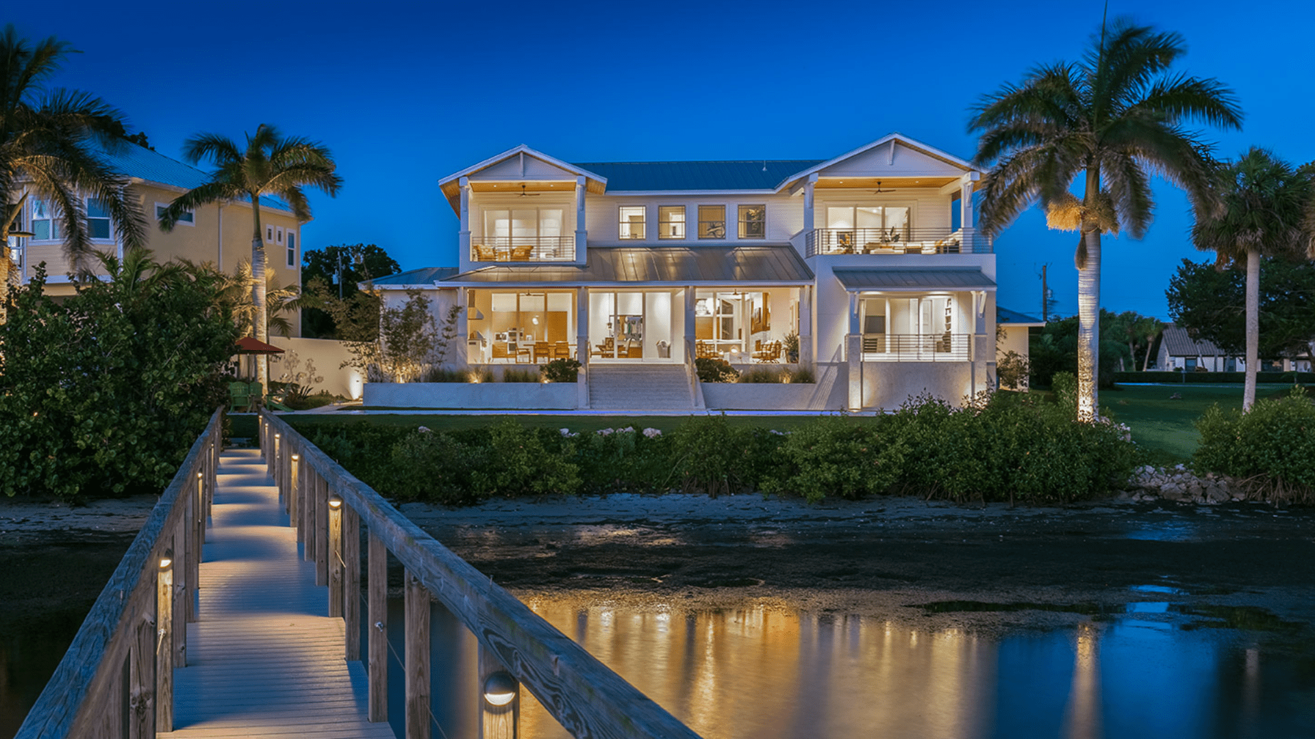 Modern, illuminated waterfront house with palm trees, captured at dusk. Wooden walkway leads over water towards the elegant two-story home.