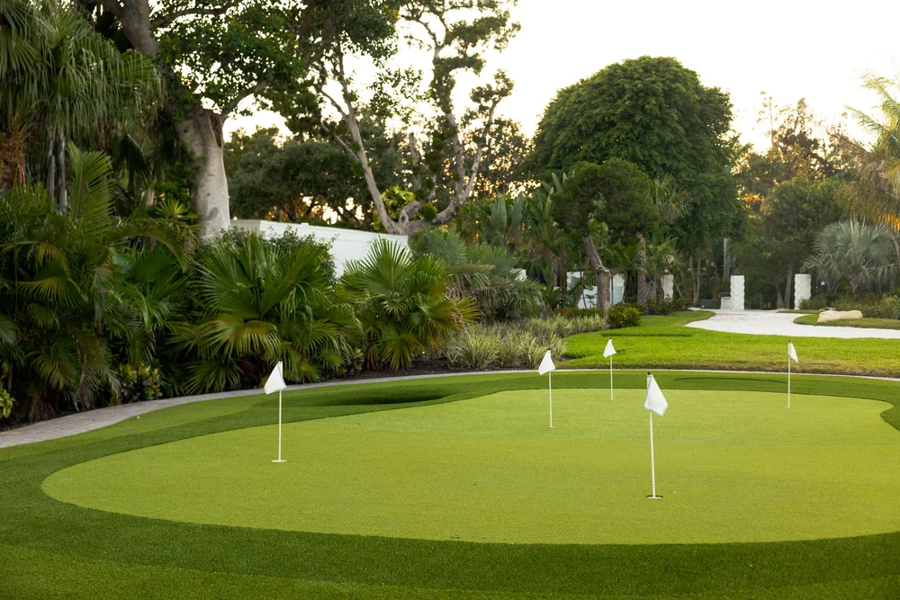 Putting green surrounded by lush tropical plants and trees, featuring five small flags, set in a peaceful, sunlit garden area.