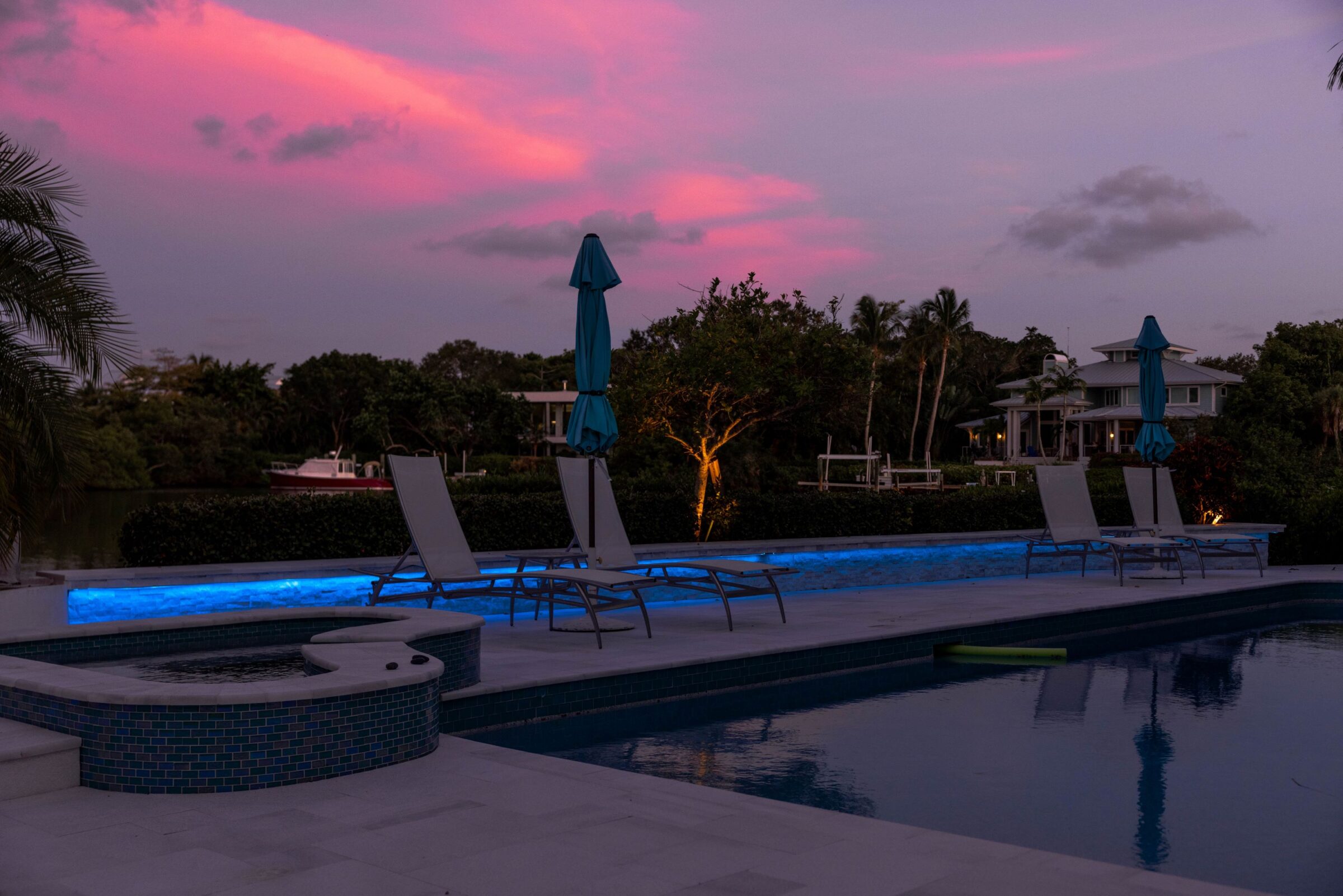 Poolside scene at sunset with lounge chairs, blue umbrellas, and palm trees. A tranquil, reflective setting under a vibrant pink sky.