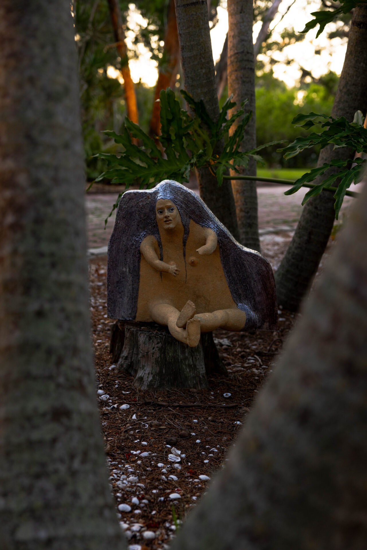 A unique person sculpture sits on a tree stump, surrounded by tall trees and scattered shells in a tranquil forest setting.
