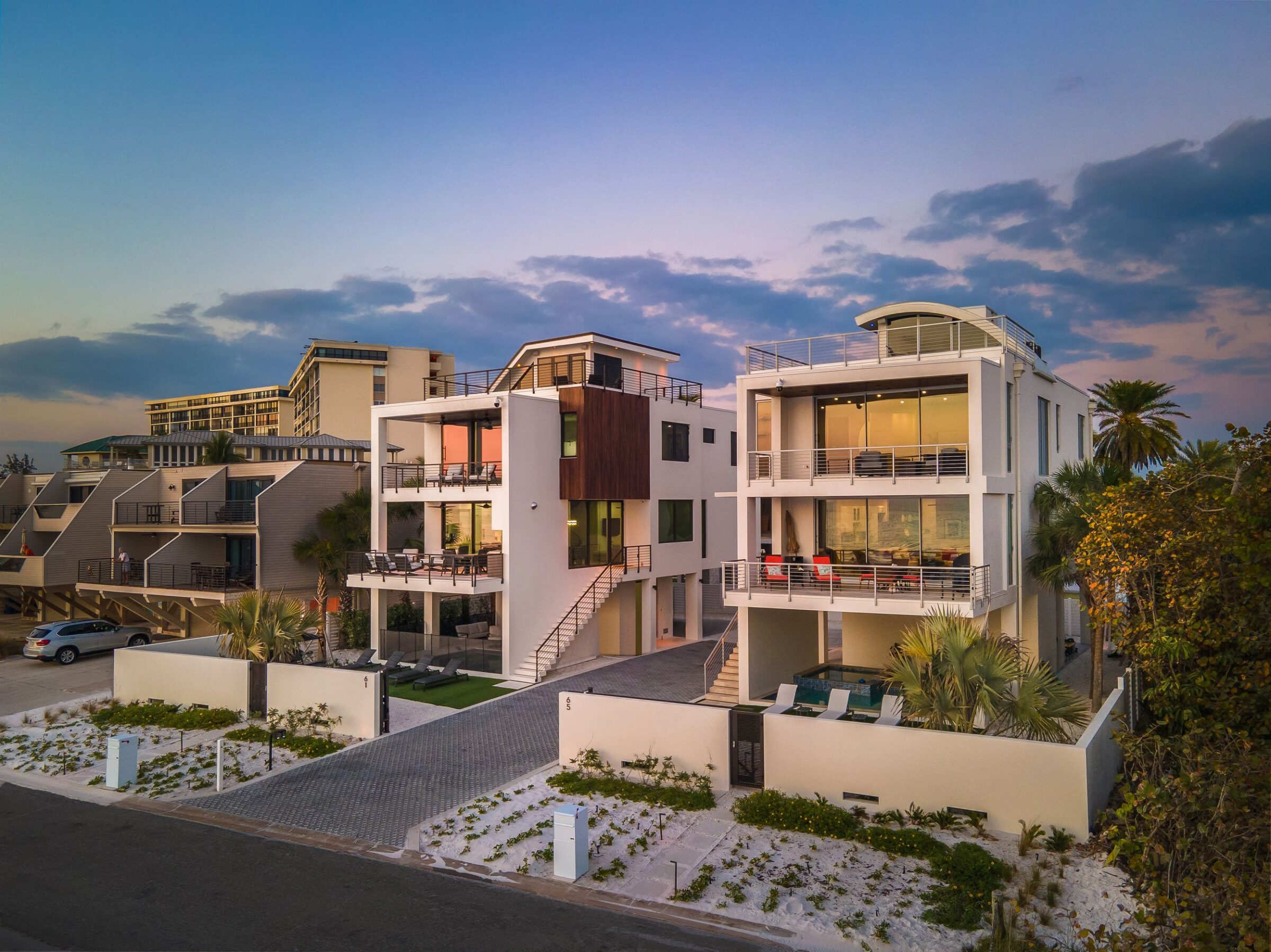Modern beachfront houses with large windows and balconies at sunset, surrounded by palm trees. A few vehicles parked on a sleek driveway.