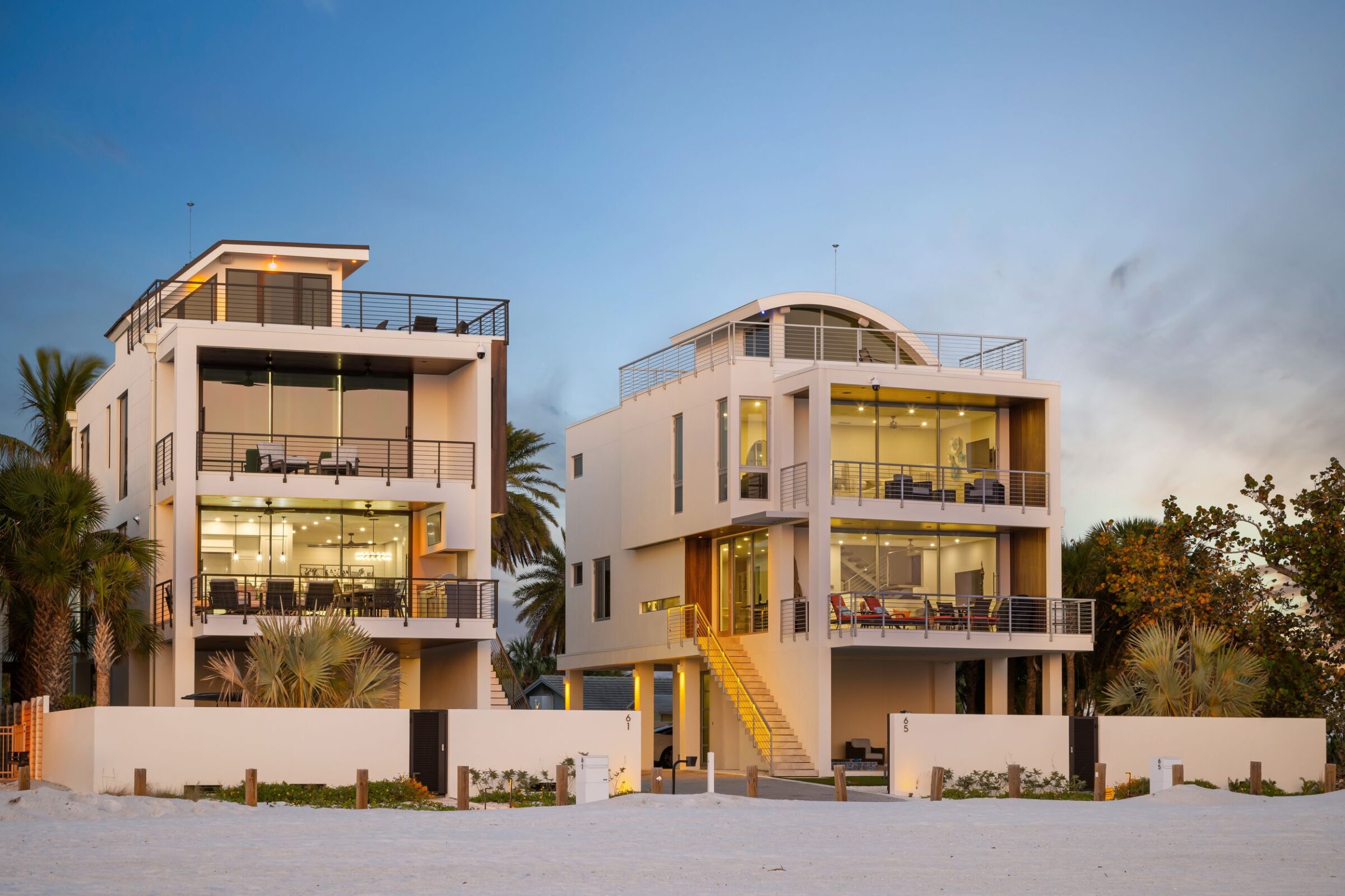 Two modern beachfront houses with expansive glass windows, surrounded by palm trees, feature rooftop terraces at dusk. No recognizable landmarks or historical buildings.