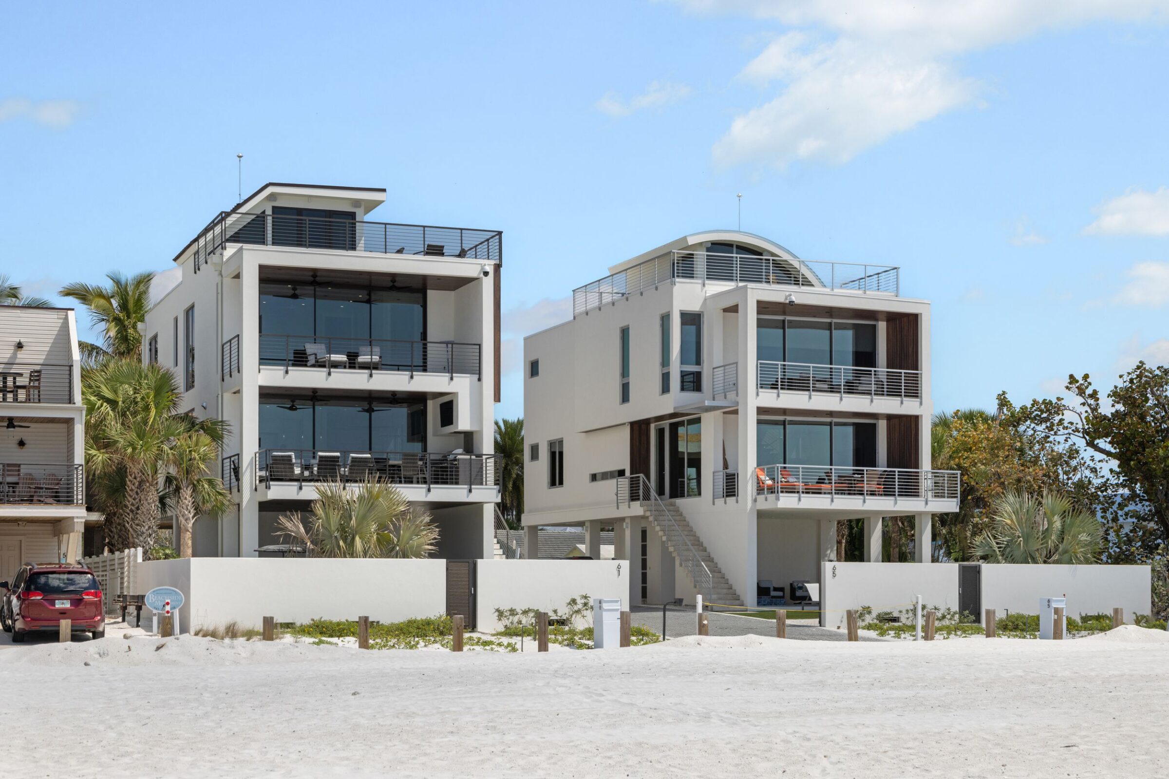 Two modern beachfront houses with large windows and balconies overlook a sandy beach. Palm trees and parked car visible nearby. Cloudy sky above.