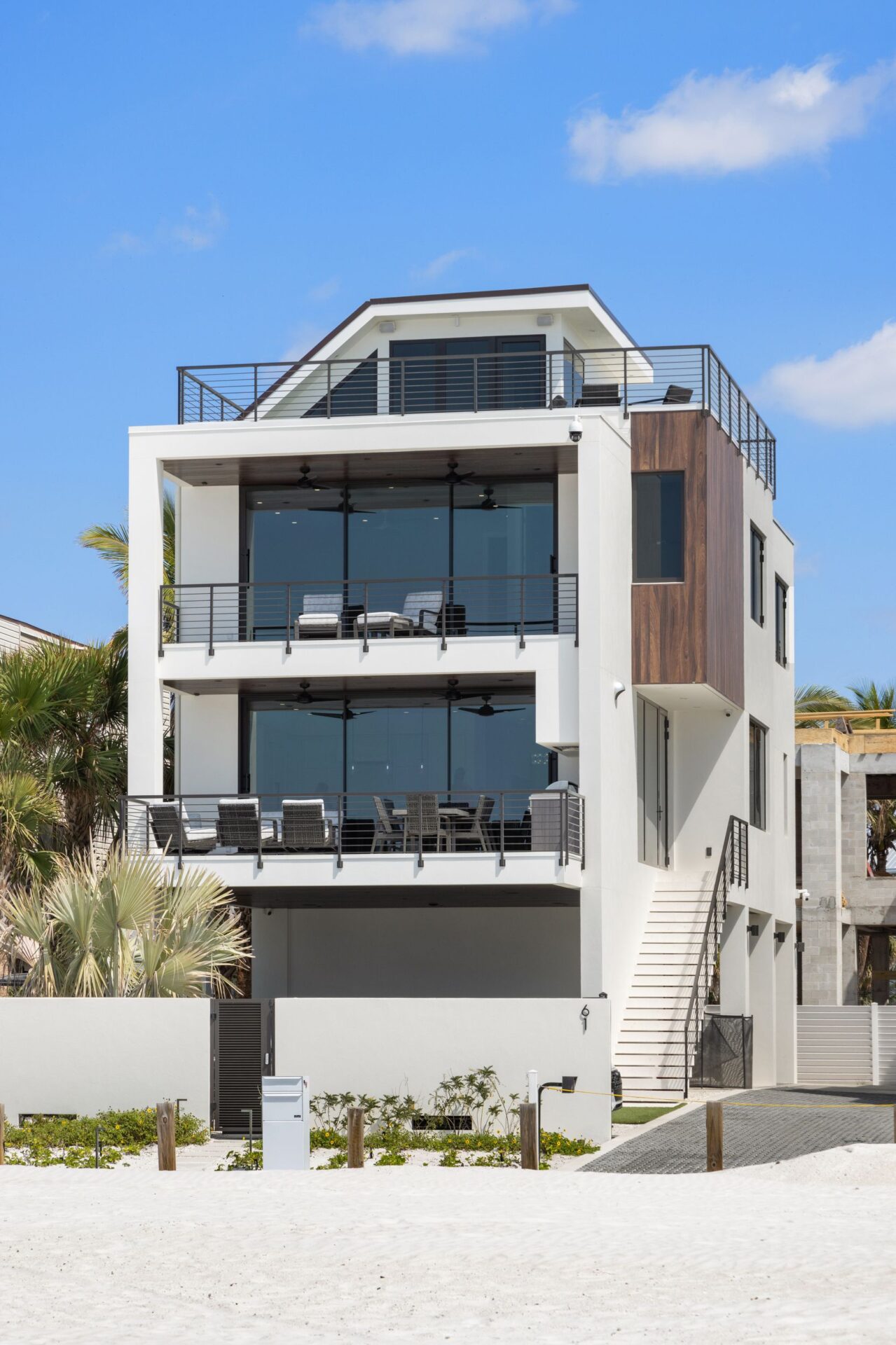 Modern beachfront house with multiple balconies, white exterior, and large windows. Surrounded by palm trees, under a bright blue sky.