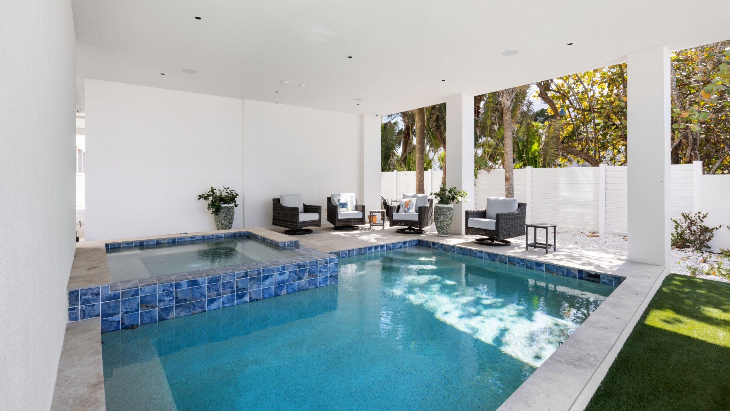 Covered patio with a small pool, blue tiles, wicker furniture, white walls, and surrounding greenery under sunlight, creating a relaxed outdoor setting.