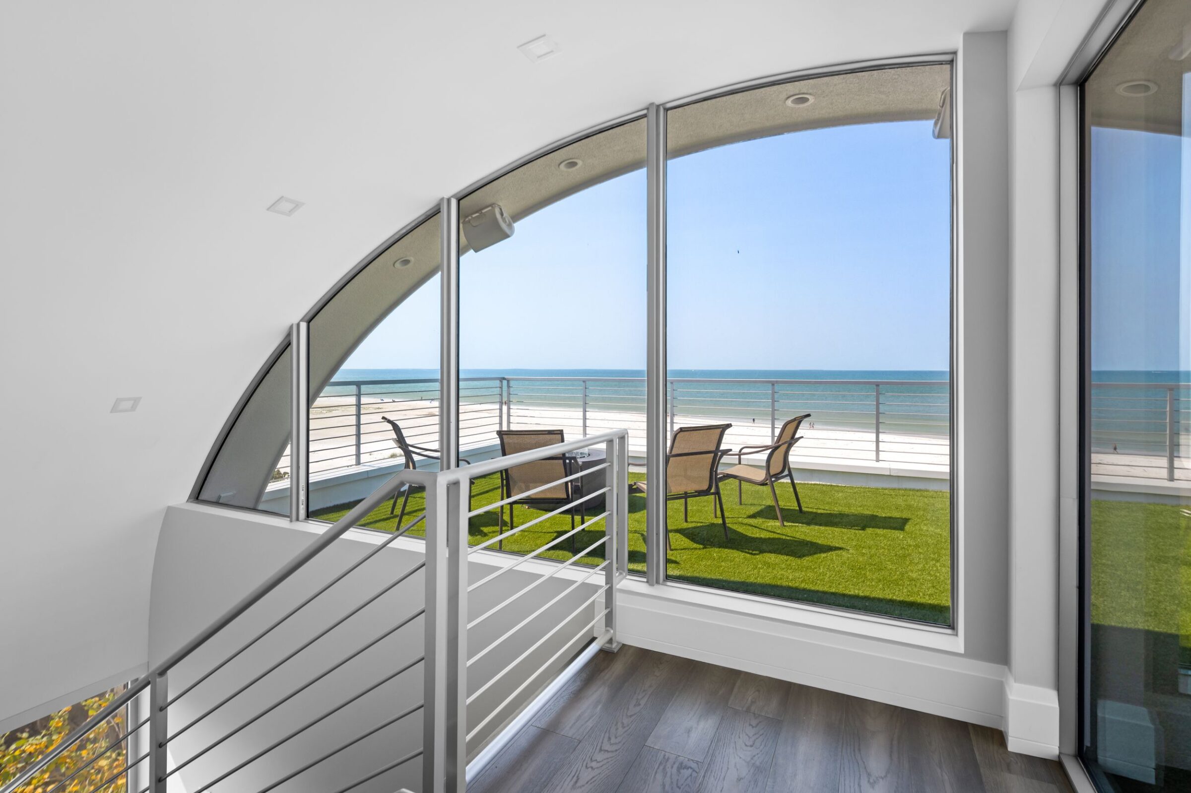 Modern interior with a large window overlooking a balcony, two chairs, and a scenic ocean view under a clear blue sky.