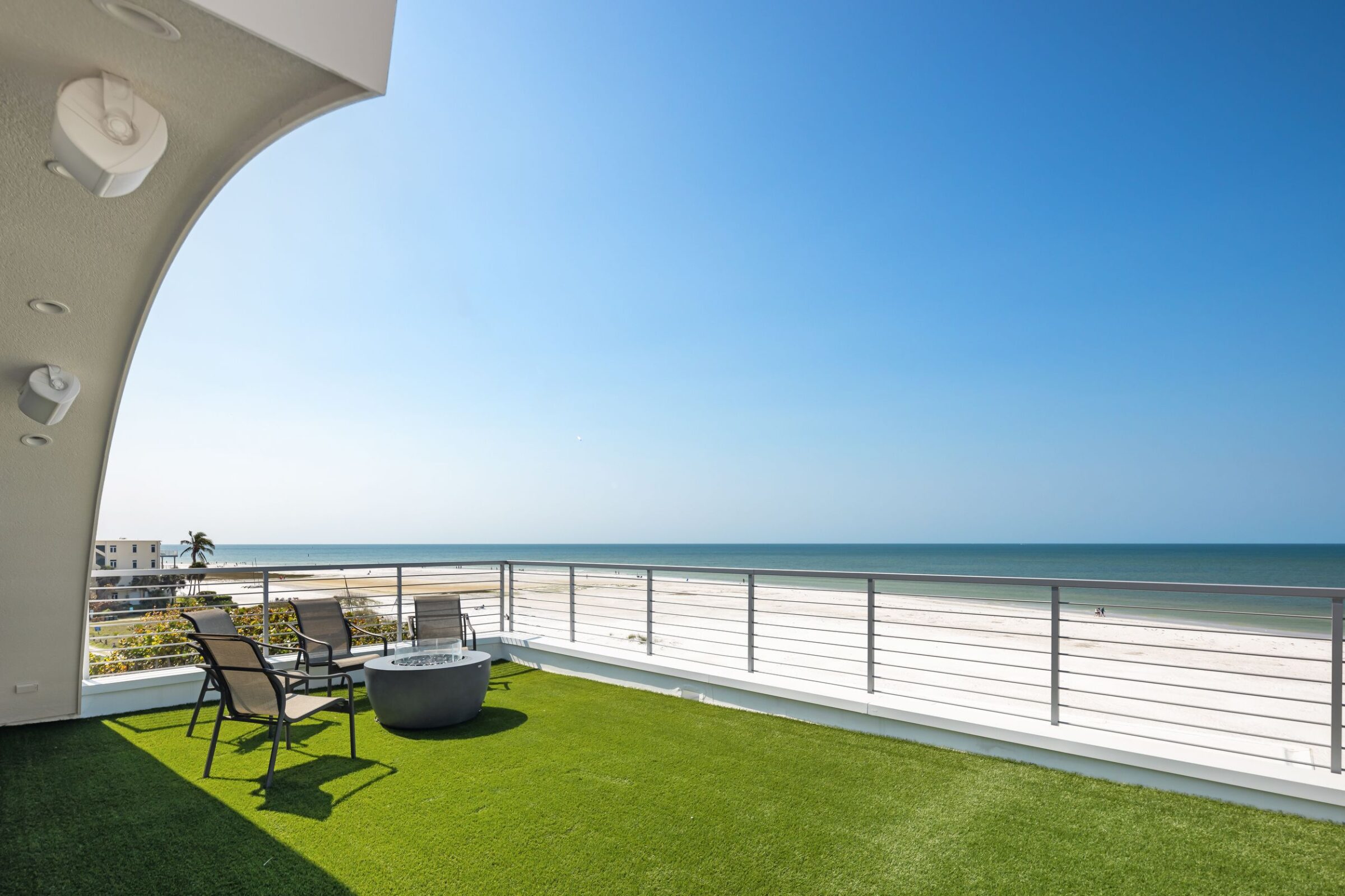 Rooftop patio with chairs and table overlooking a sandy beach and ocean under a clear blue sky, featuring a modern railing.