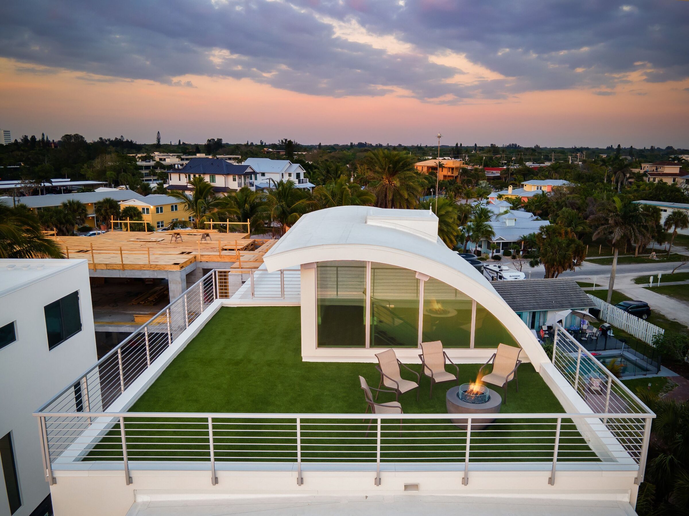 Rooftop with artificial grass, chairs, and fire pit; overlooking a neighborhood with palm trees and houses at sunset. No landmarks visible.