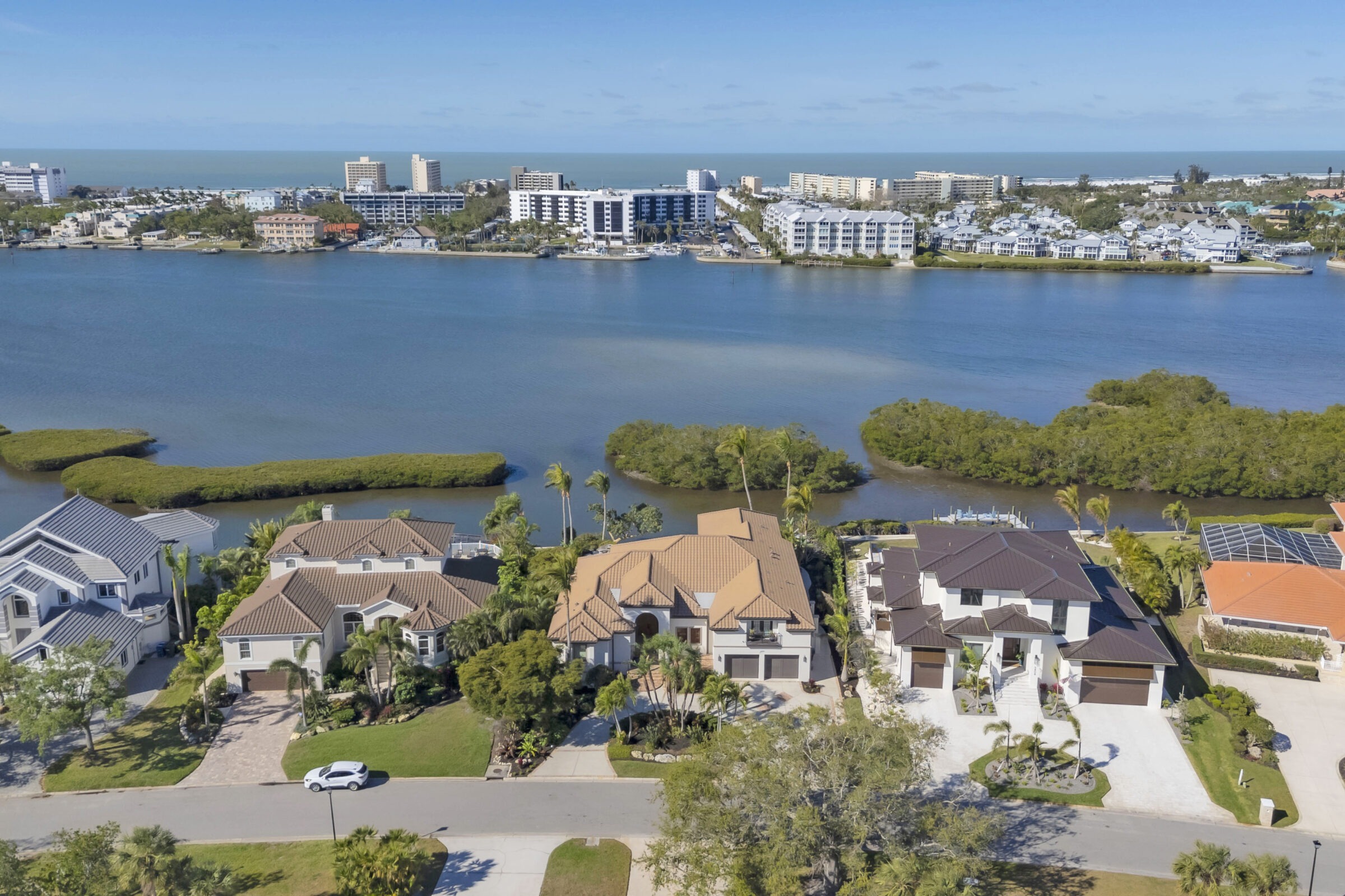 Coastal landscape features waterfront homes, lush greenery, and a calm bay with distant modern buildings, under a clear blue sky.