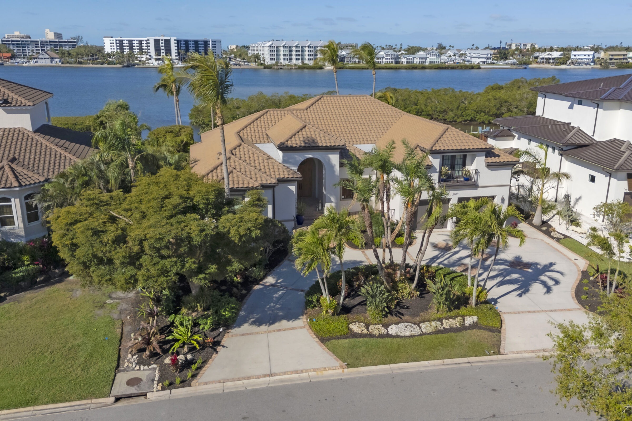 Waterfront home with palm trees and a curved driveway, overlooking buildings across a body of water. Sunny day with clear skies.