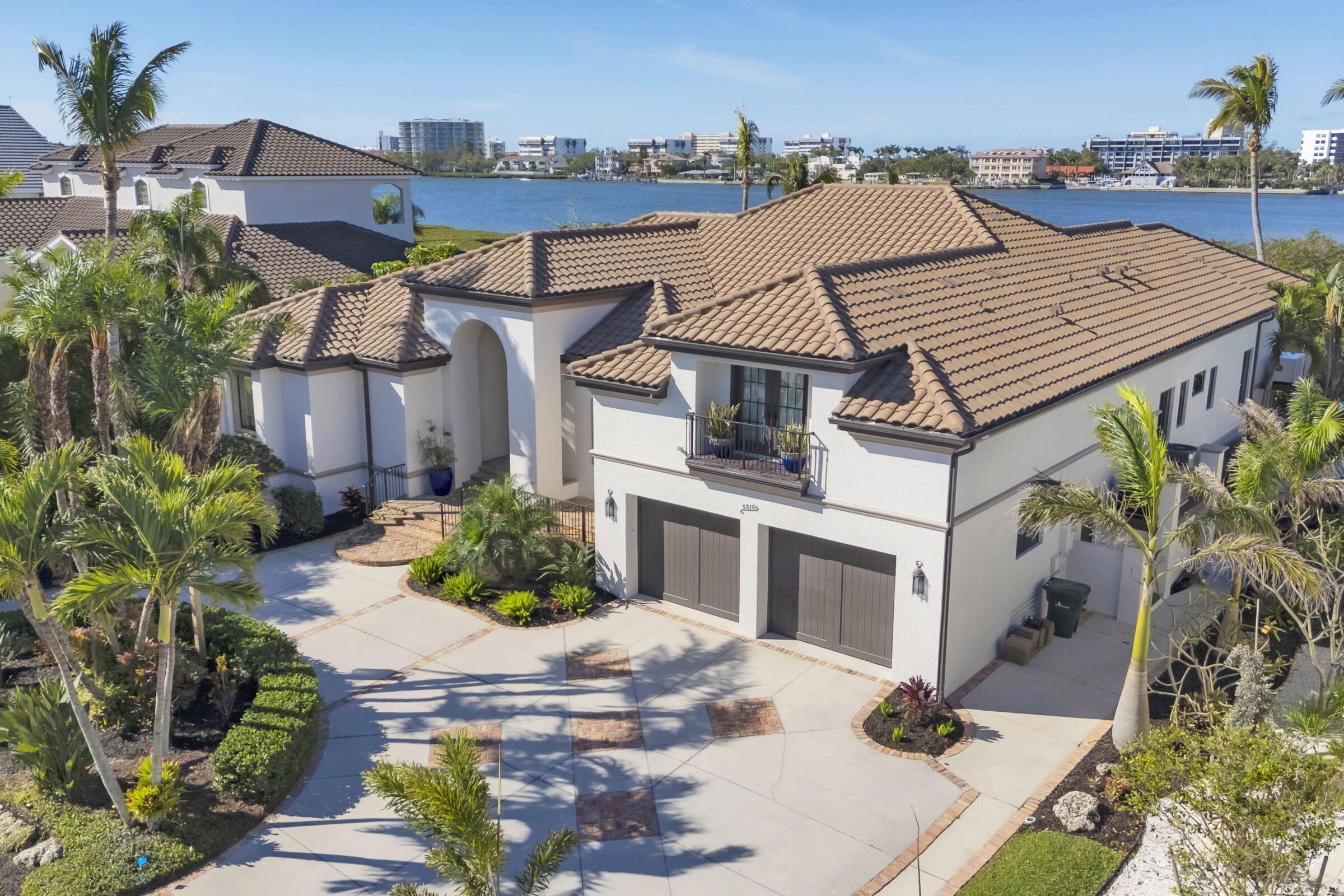 Elegant waterfront home with a tile roof and landscaping, overlooking a serene lake. Modern buildings visible in the distant skyline.