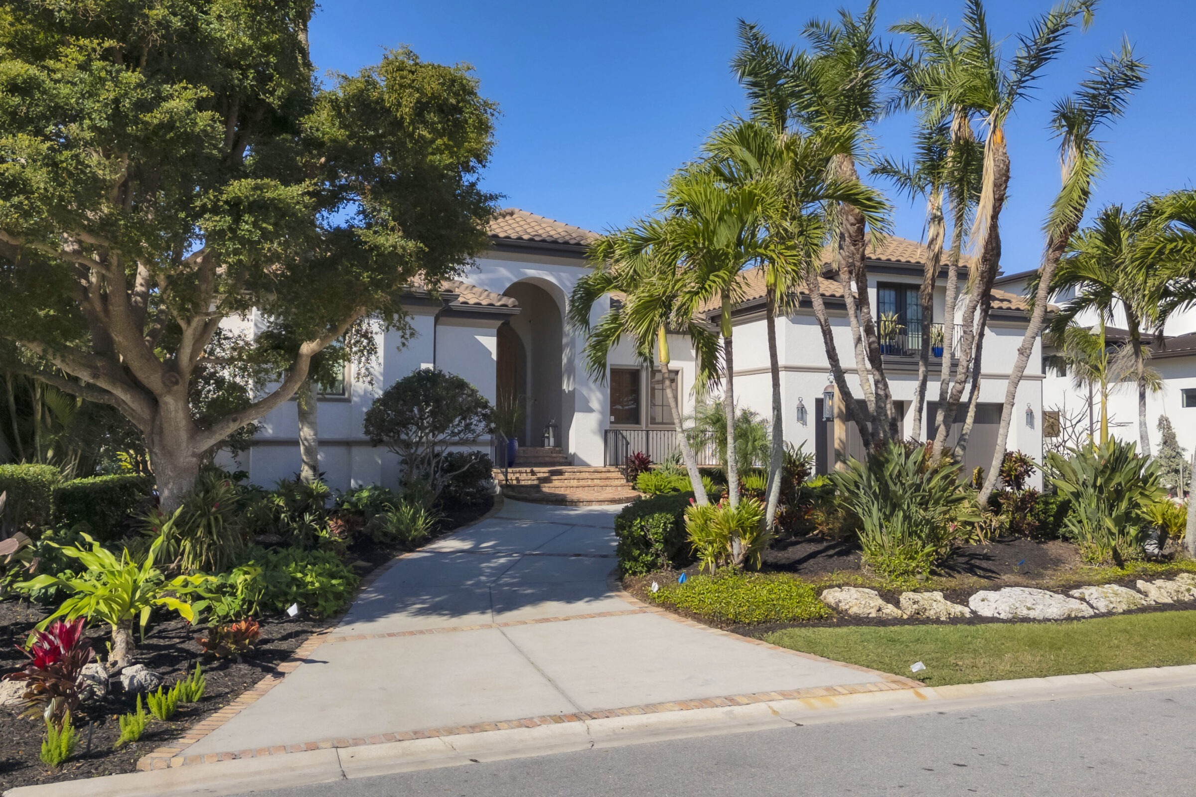 Modern house with arched entrance, terracotta roof, surrounded by lush tropical landscaping and palm trees, under a clear blue sky. No people visible.