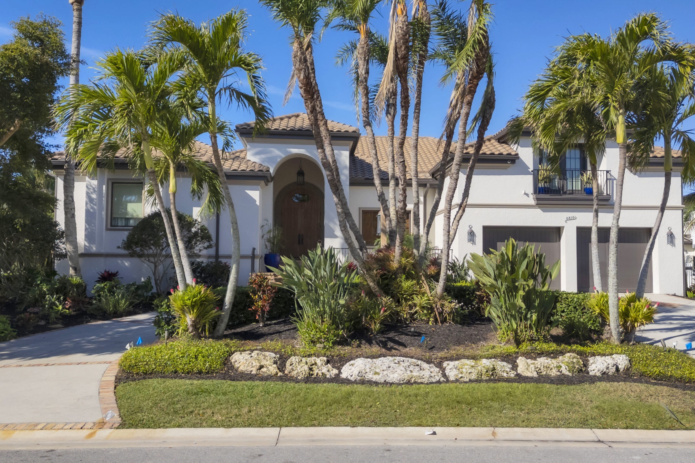 A modern house with a tiled roof, surrounded by palm trees and lush landscaping, under a clear blue sky with no people visible.