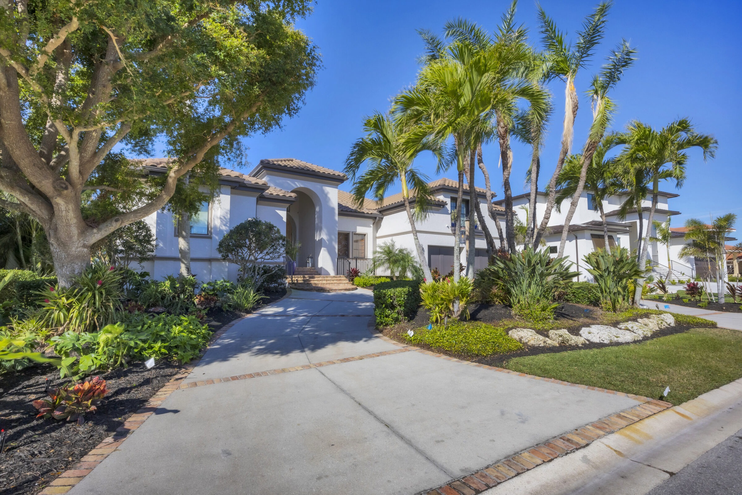 A modern house with Mediterranean architecture, surrounded by lush greenery and palm trees, under a bright blue sky. No landmarks visible.