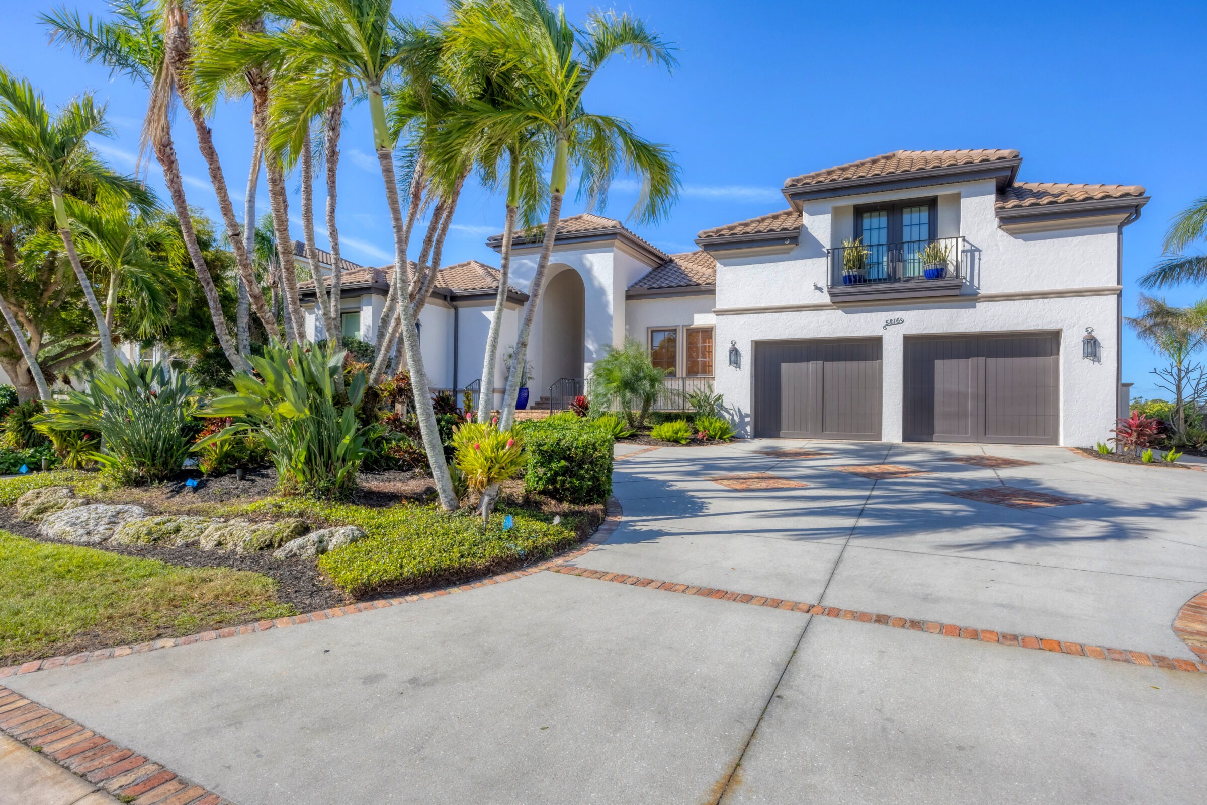 A Mediterranean-style house with palm trees, manicured garden, and spacious driveway under a clear blue sky. No people present, vibrant landscaping.