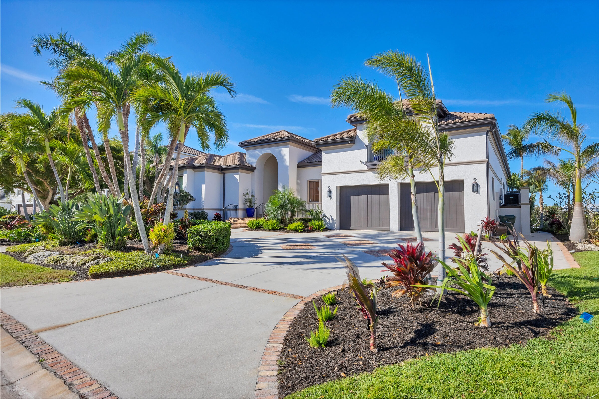 A modern house with palm trees and landscaped garden under a clear blue sky, featuring a three-car garage and tiled roof.