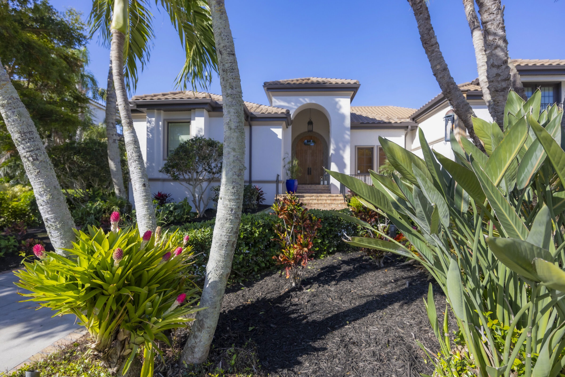 A Mediterranean-style house with a tiled roof and archway, surrounded by tropical plants and trees under a clear blue sky.