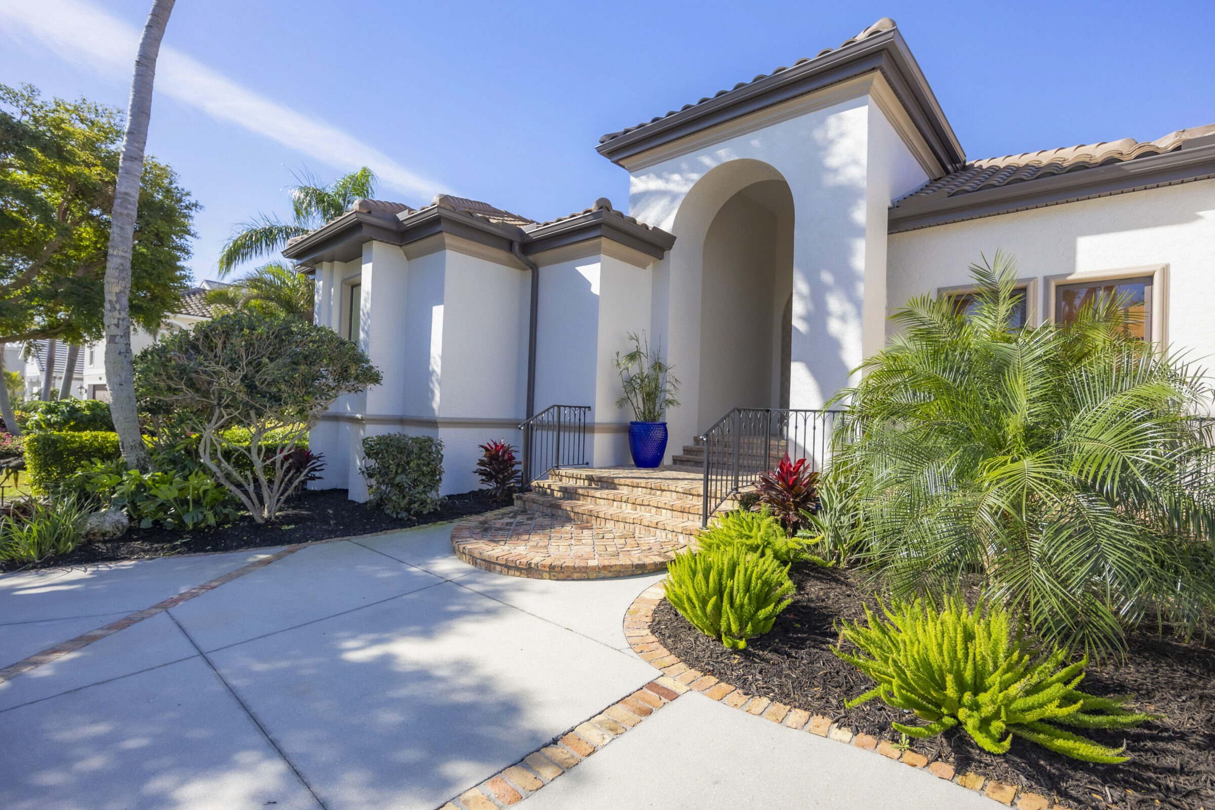 A modern white house with a tiled roof, surrounded by lush greenery and a manicured garden path under a clear blue sky.