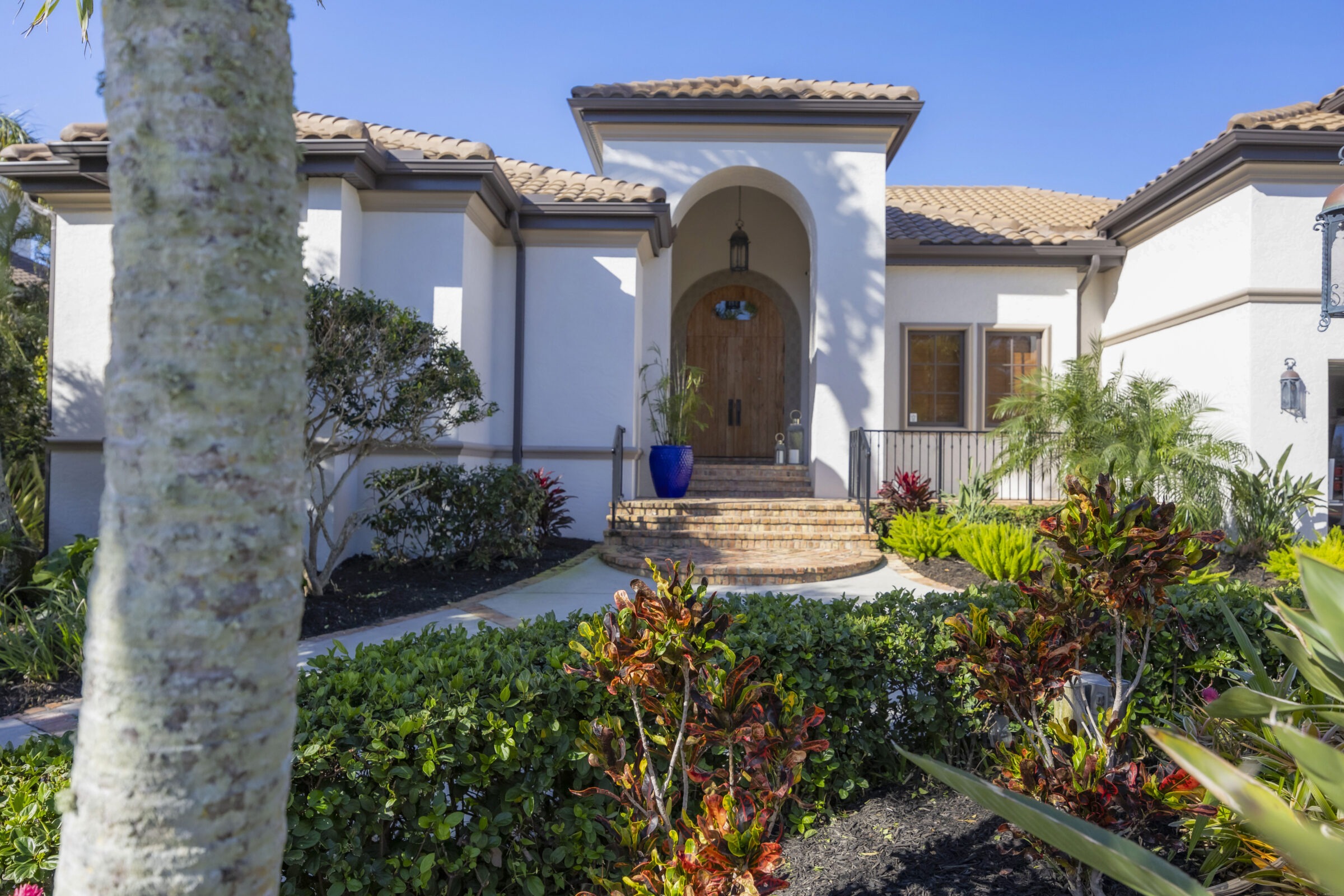 A Mediterranean-style house with an arched entrance, surrounded by lush greenery and tropical plants, under a clear blue sky.
