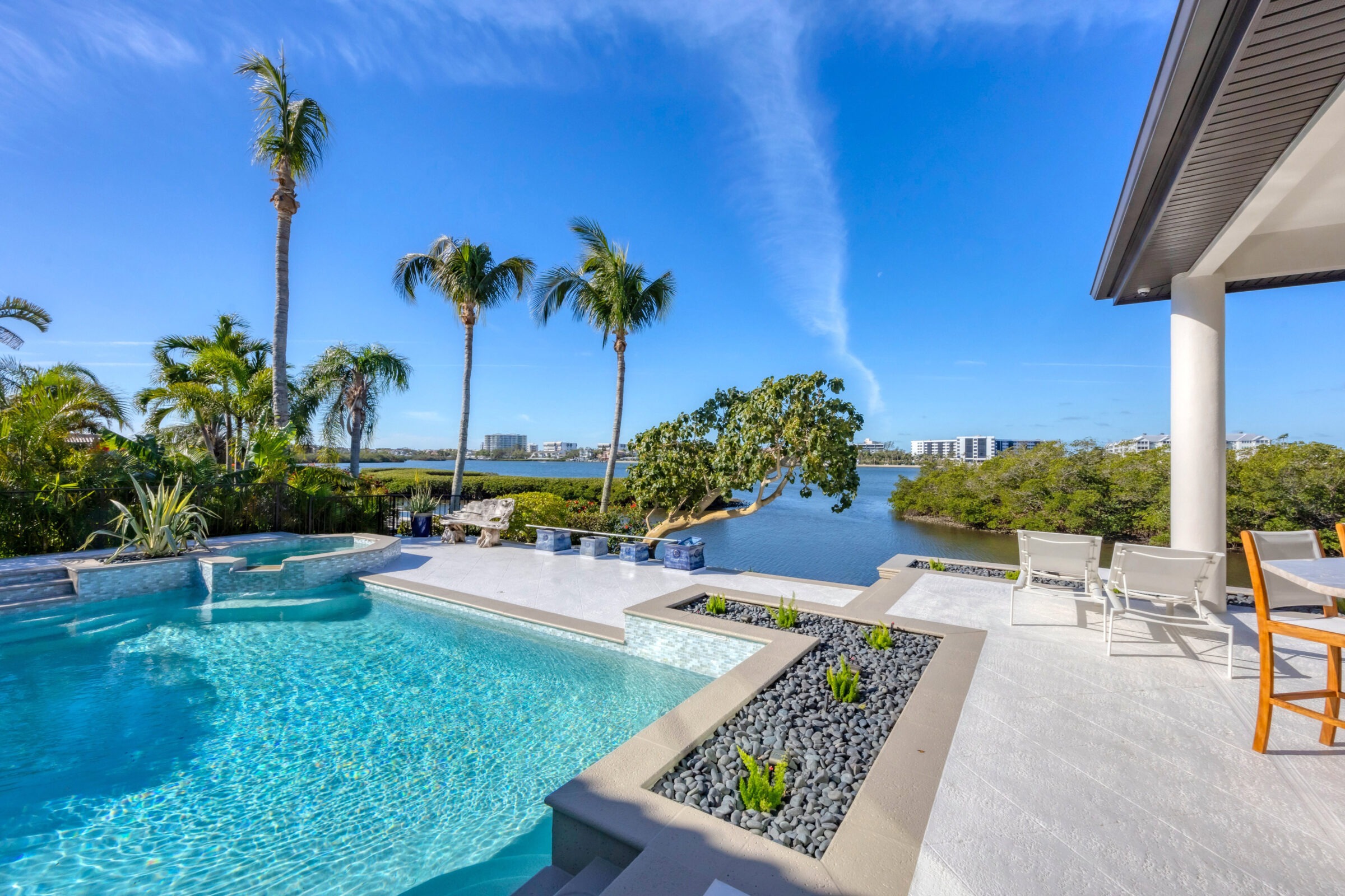 Poolside area with palm trees overlooks a serene river, modern buildings in the background, and clear blue sky above.