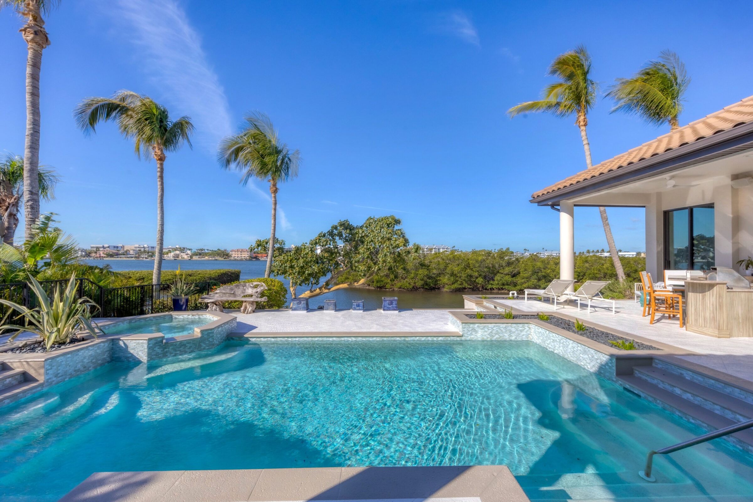 Luxurious poolside oasis with palm trees, overlooking a serene waterway. Modern patio furniture and clear blue skies complete the idyllic coastal setting.