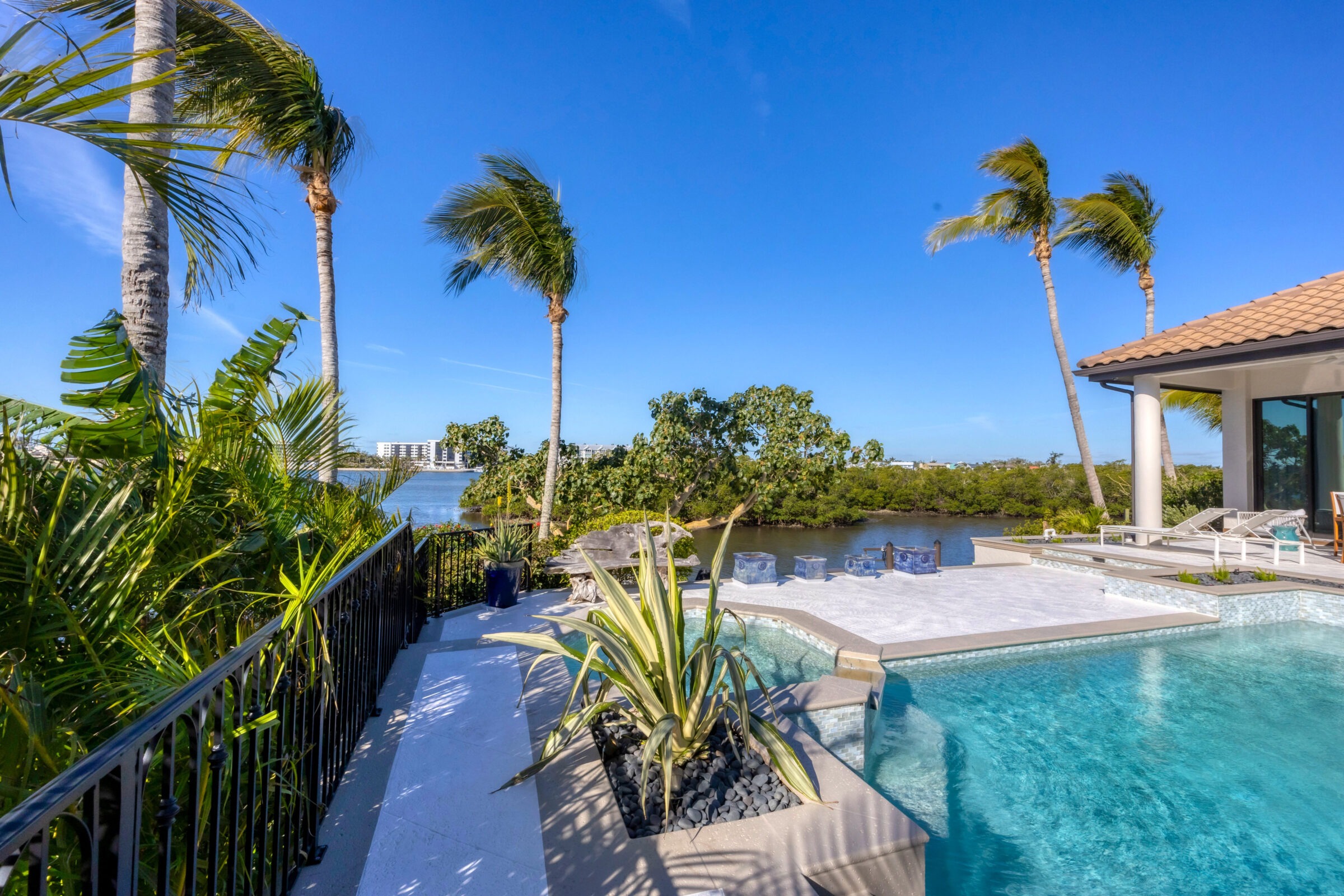 Poolside patio with palm trees overlooking a waterway and distant buildings under a clear blue sky.