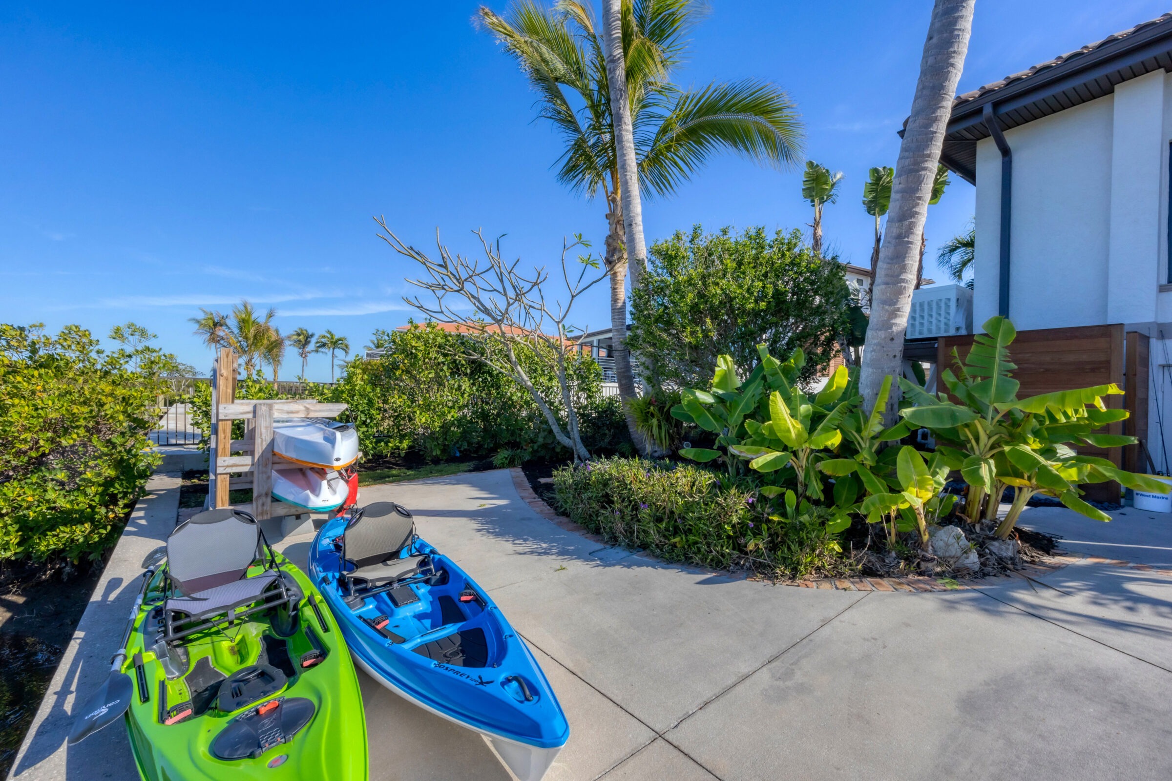 Colorful kayaks rest on a concrete path surrounded by lush tropical plants and clear blue skies in a serene, idyllic setting.