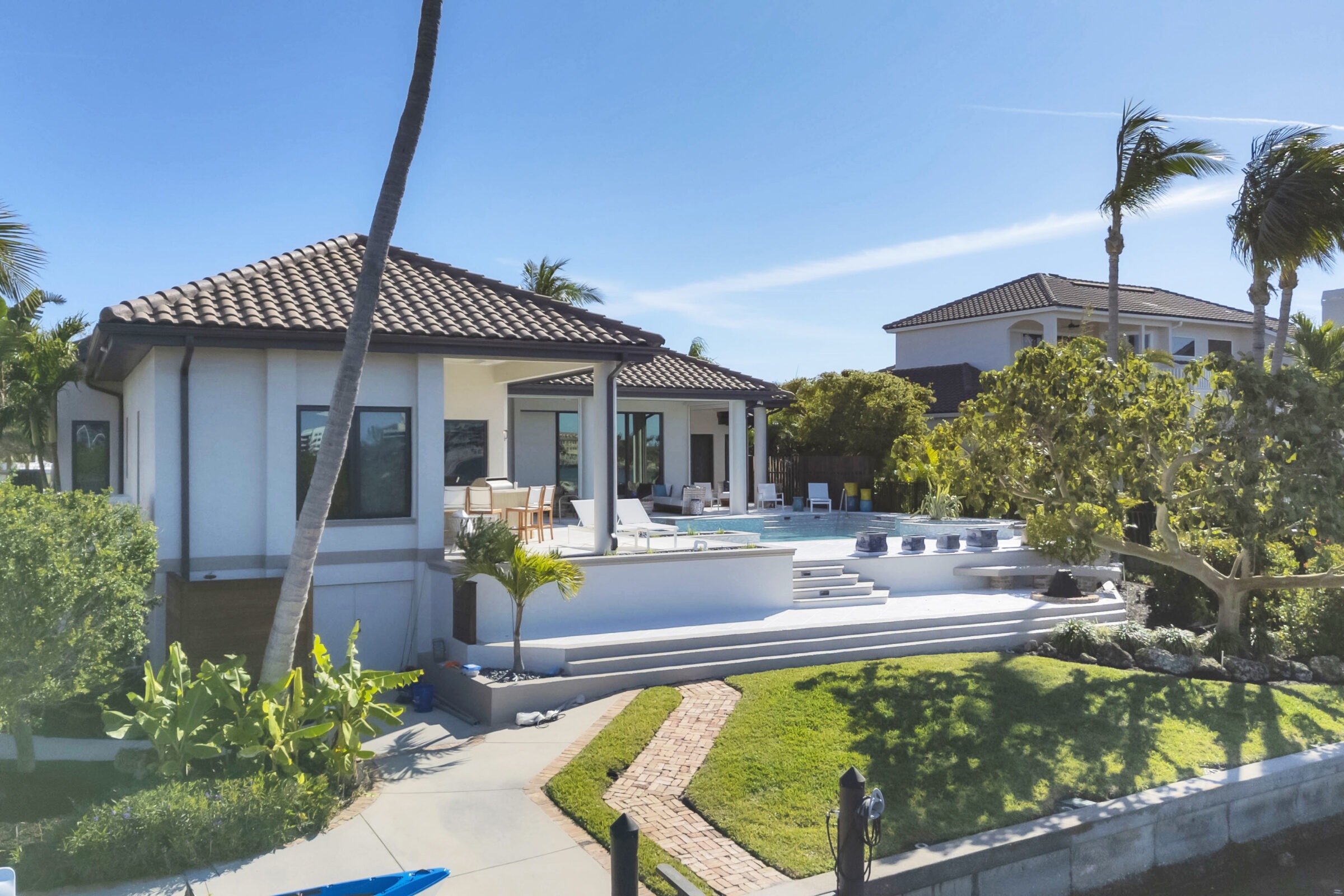 Modern white house with a pool, surrounded by palm trees and lush greenery, under a clear blue sky. Steps lead to garden.