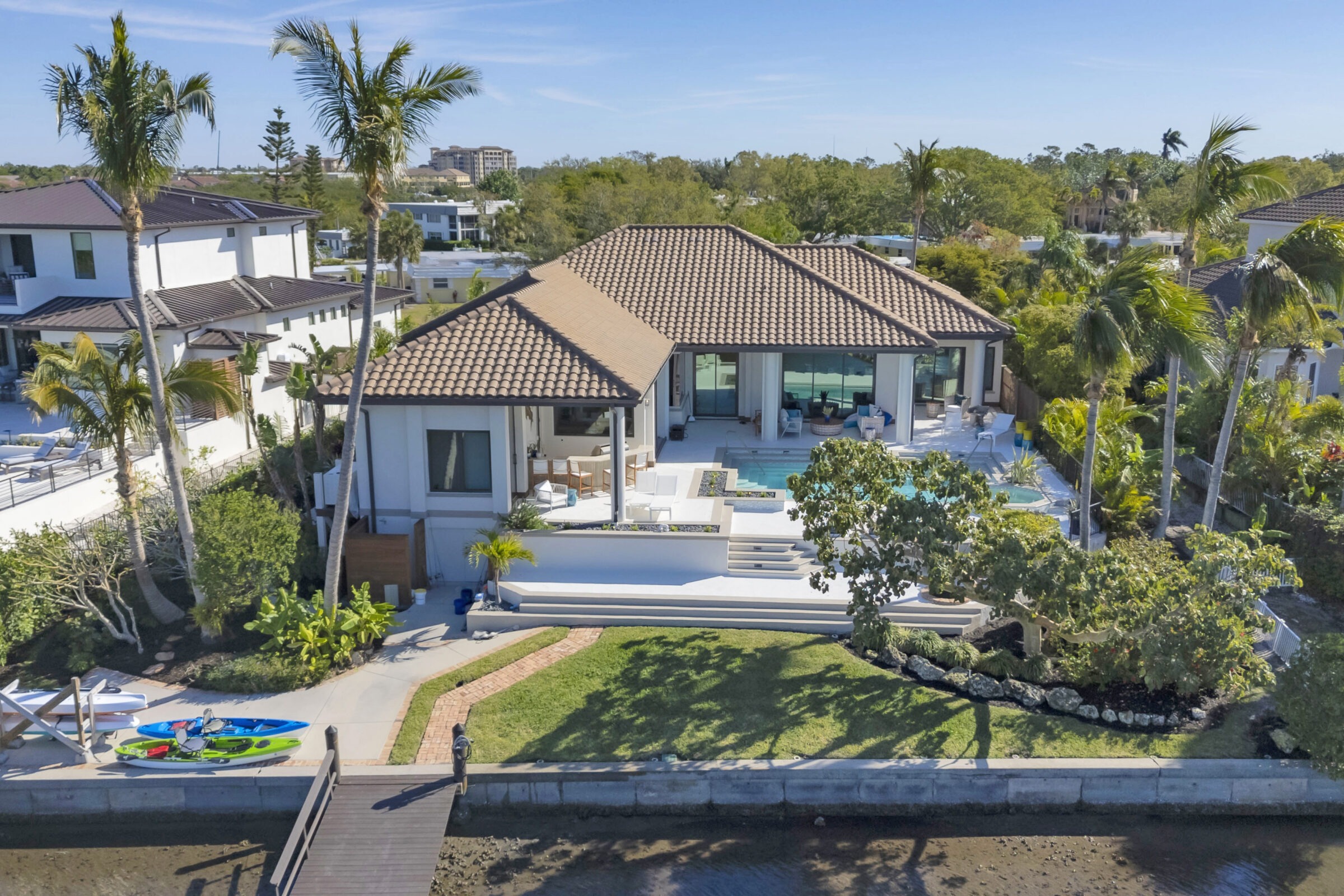 A modern waterfront house with a tiled roof, pool, patio, palm trees, canoes, and a wooden dock, surrounded by tropical greenery.