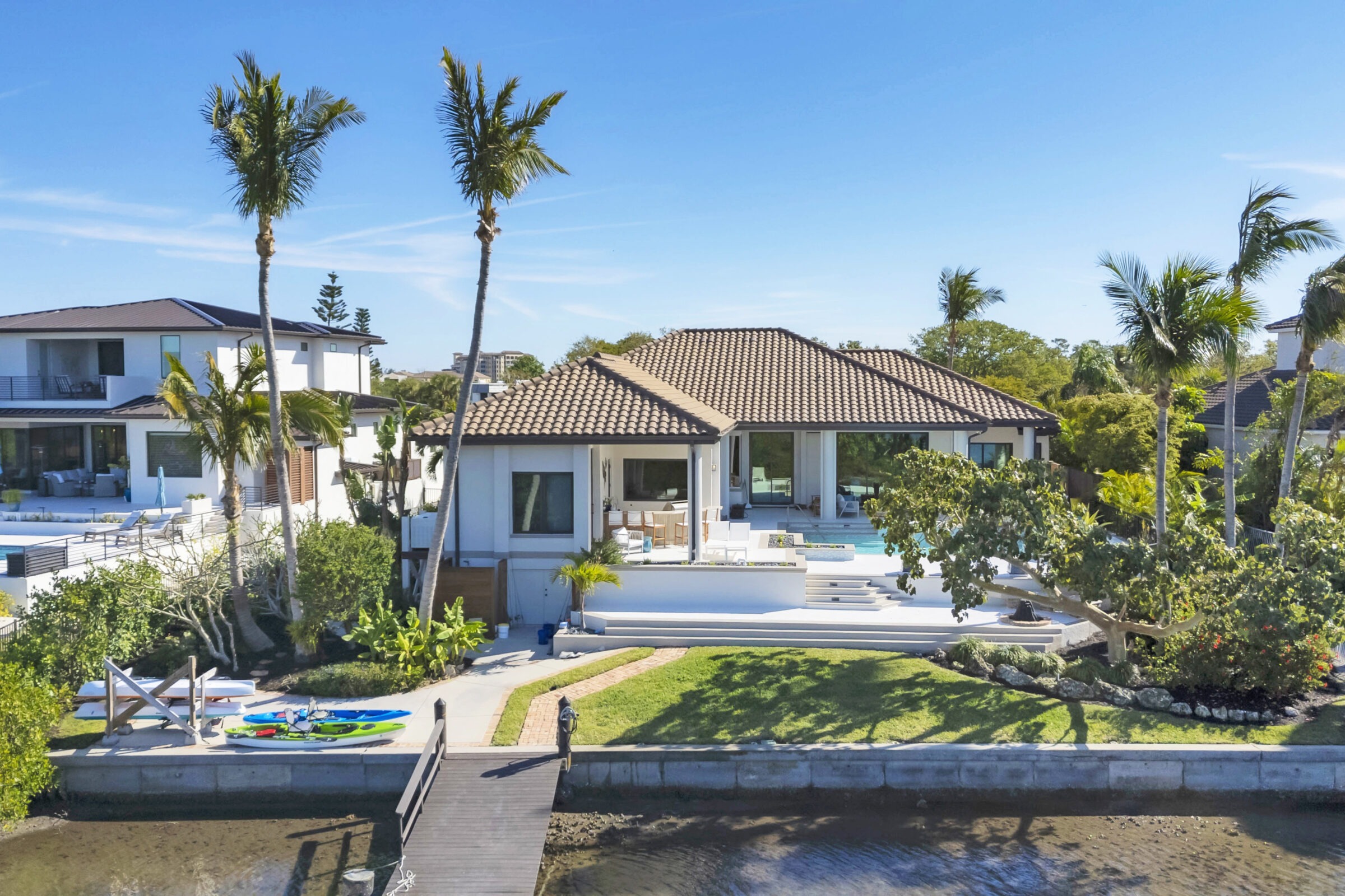 Modern waterfront house with palm trees, dock, and kayaks. Spacious patio and swimming pool, surrounded by greenery under a clear, blue sky.