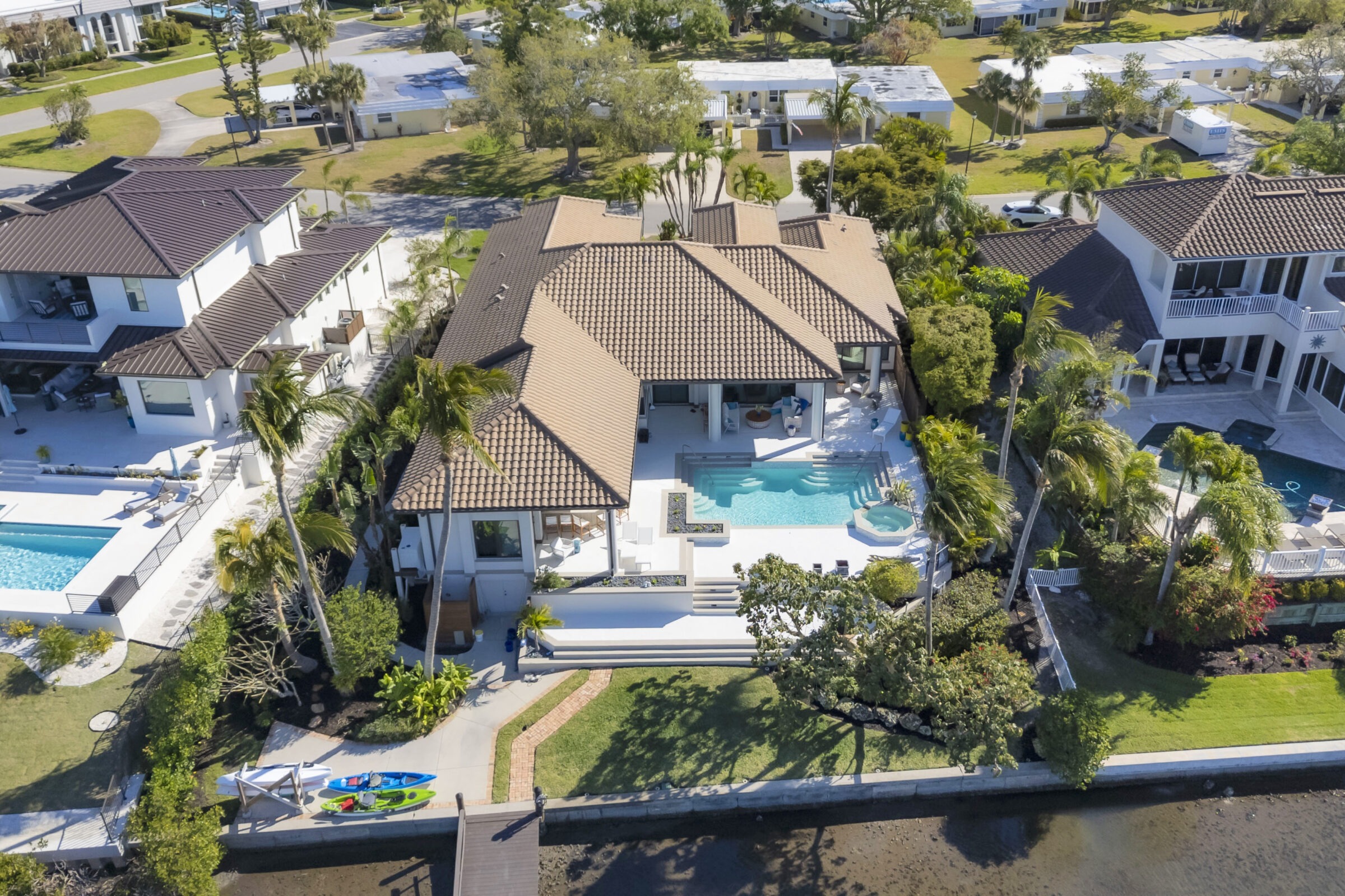 Luxurious waterfront home with a large pool and lush palm trees. Nearby properties feature modern designs, surrounded by a peaceful residential neighborhood.