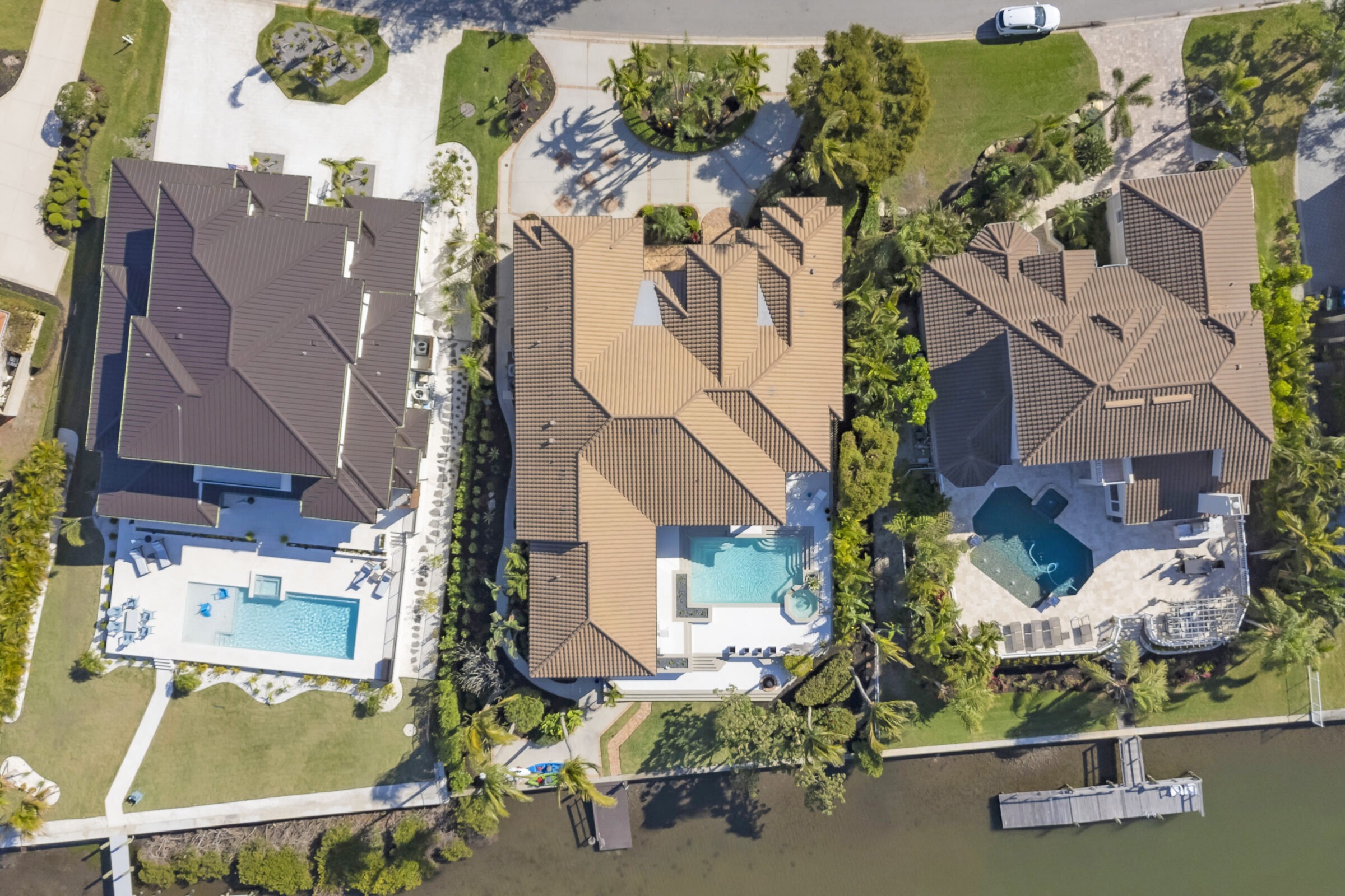 Aerial view of three luxurious waterfront homes with pools in a residential area; features greenery and a road with a parked car.
