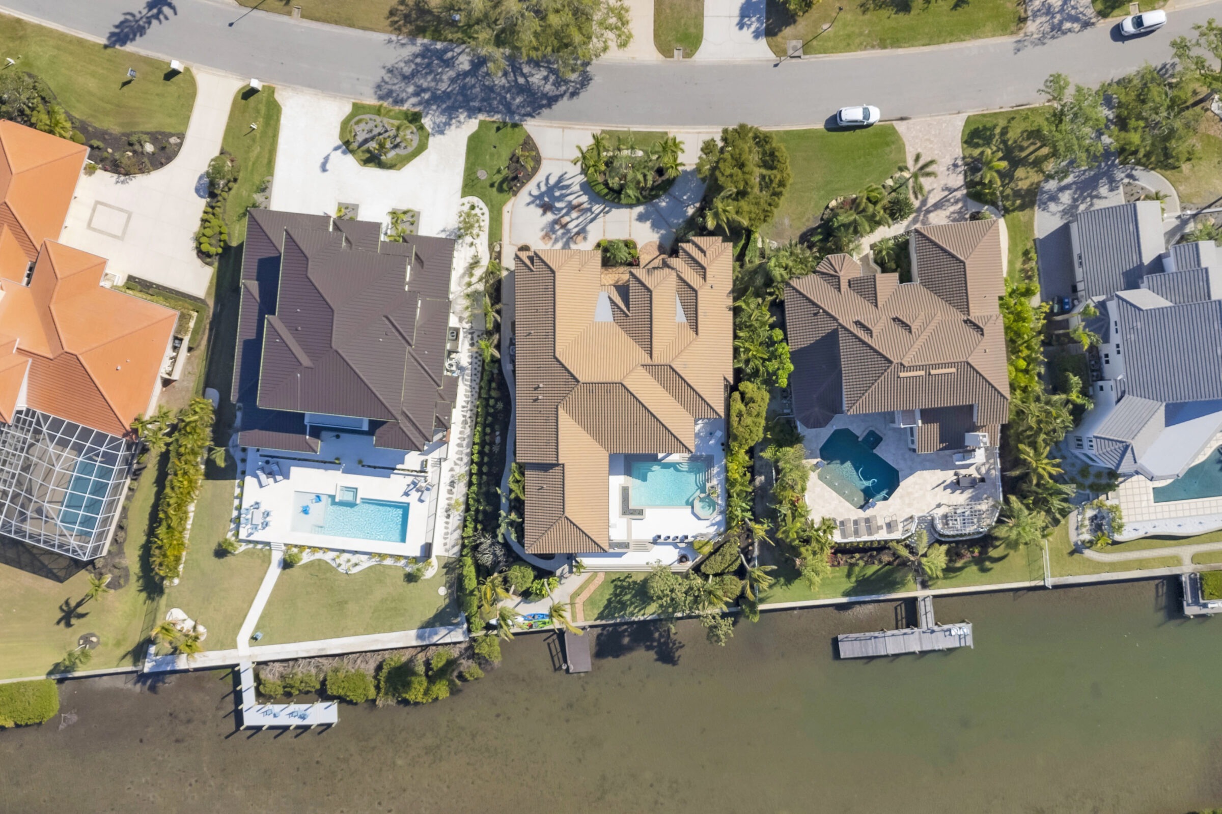 Aerial view of waterfront homes with pools, neatly landscaped gardens, and private docks along a tranquil canal, bordered by a quiet street.