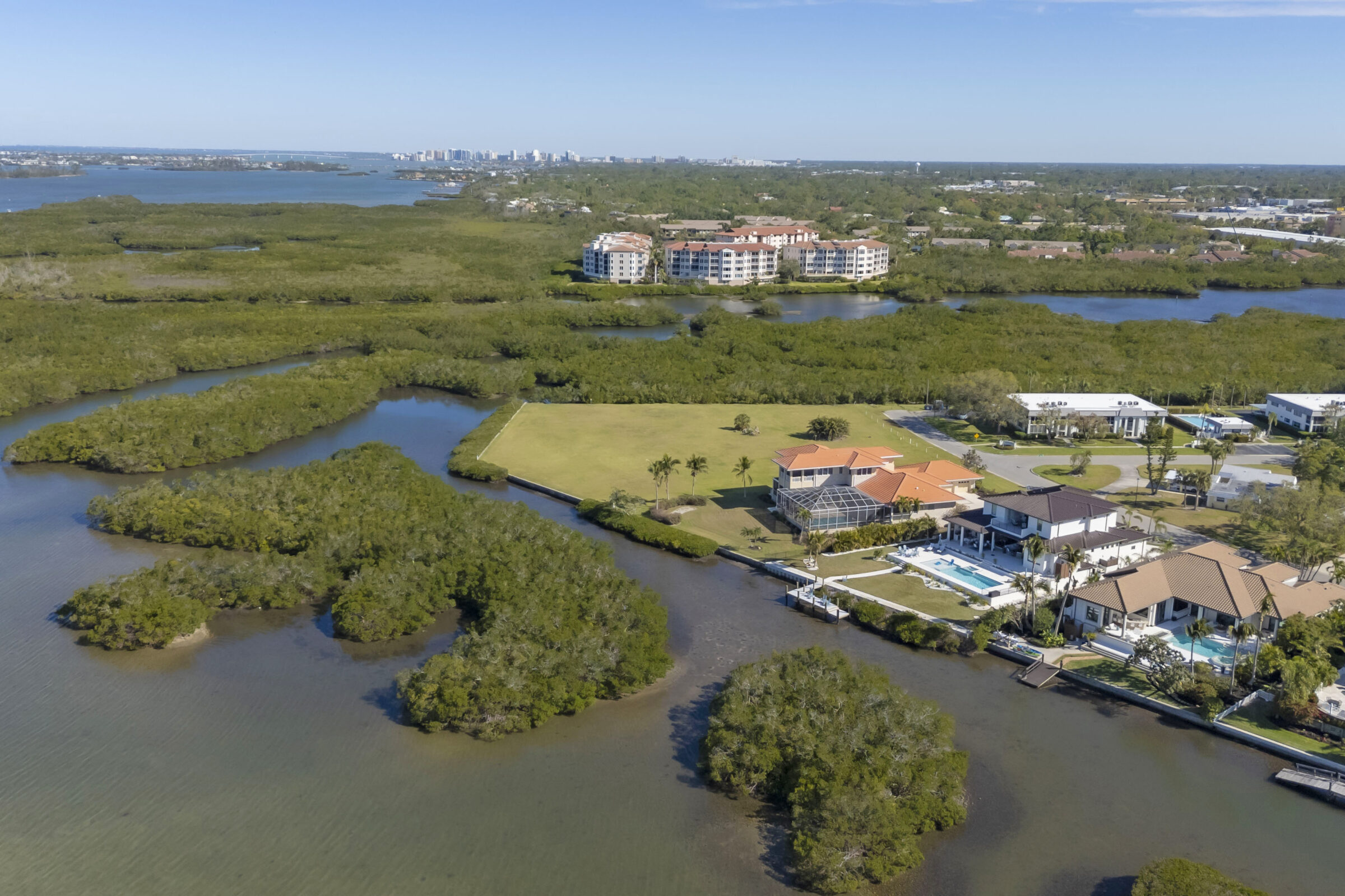 Aerial view of luxurious waterfront homes with pools, surrounded by lush mangroves and water channels, with a cityscape visible in the distance.