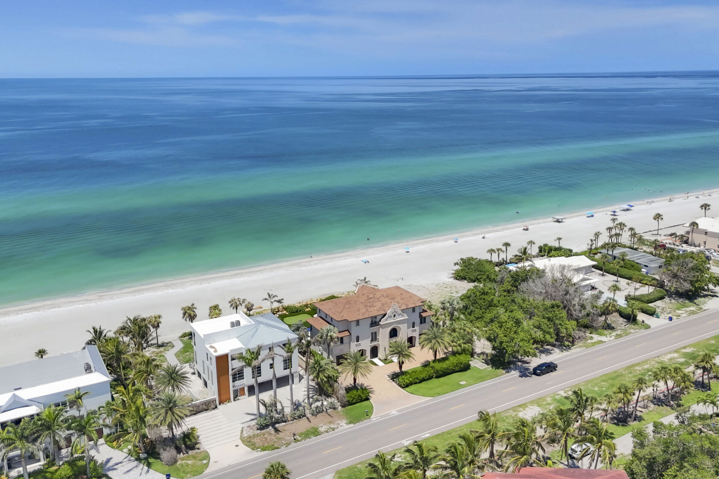 Coastal scene with luxury beachfront homes, palm trees, a sandy beach, and calm turquoise ocean waves under a clear blue sky.