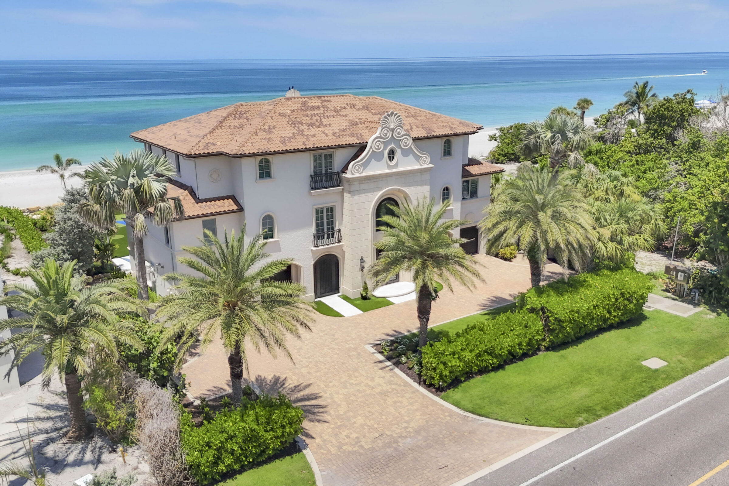 Elegant beachfront villa with terracotta roof, lush palm trees, and ocean view; expansive entrance and driveway leading to turquoise waters, clear skies above.