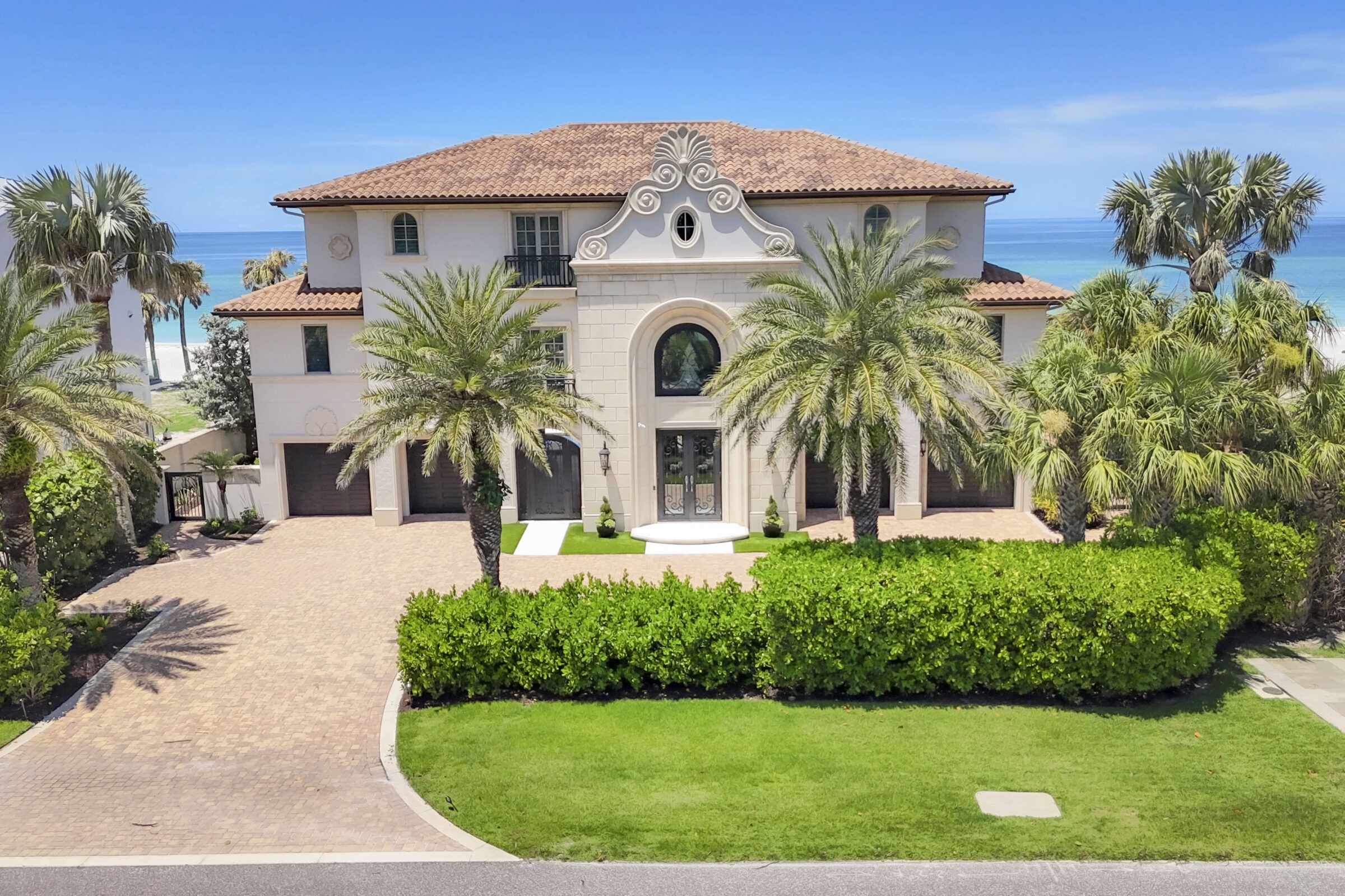 A Mediterranean-style beachfront villa with palm trees, lush landscaping, and an ocean backdrop visible beyond the property, under a clear blue sky.