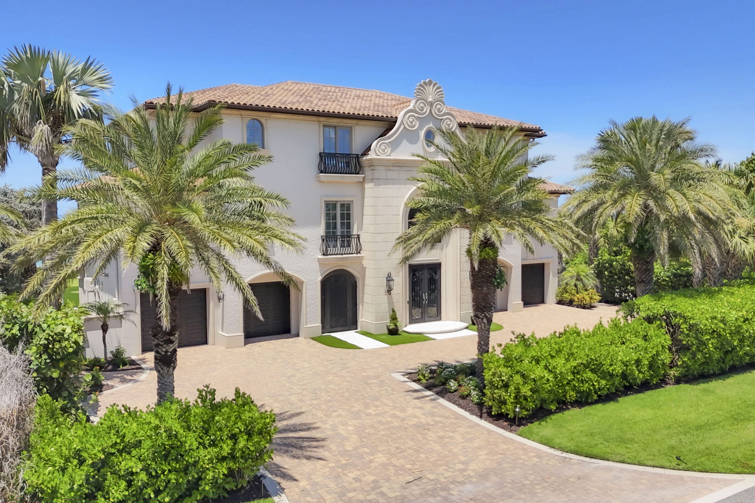 Elegant Mediterranean-style house with arched windows, palm trees, and a paved driveway. Lush greenery surrounds the property under a clear blue sky.