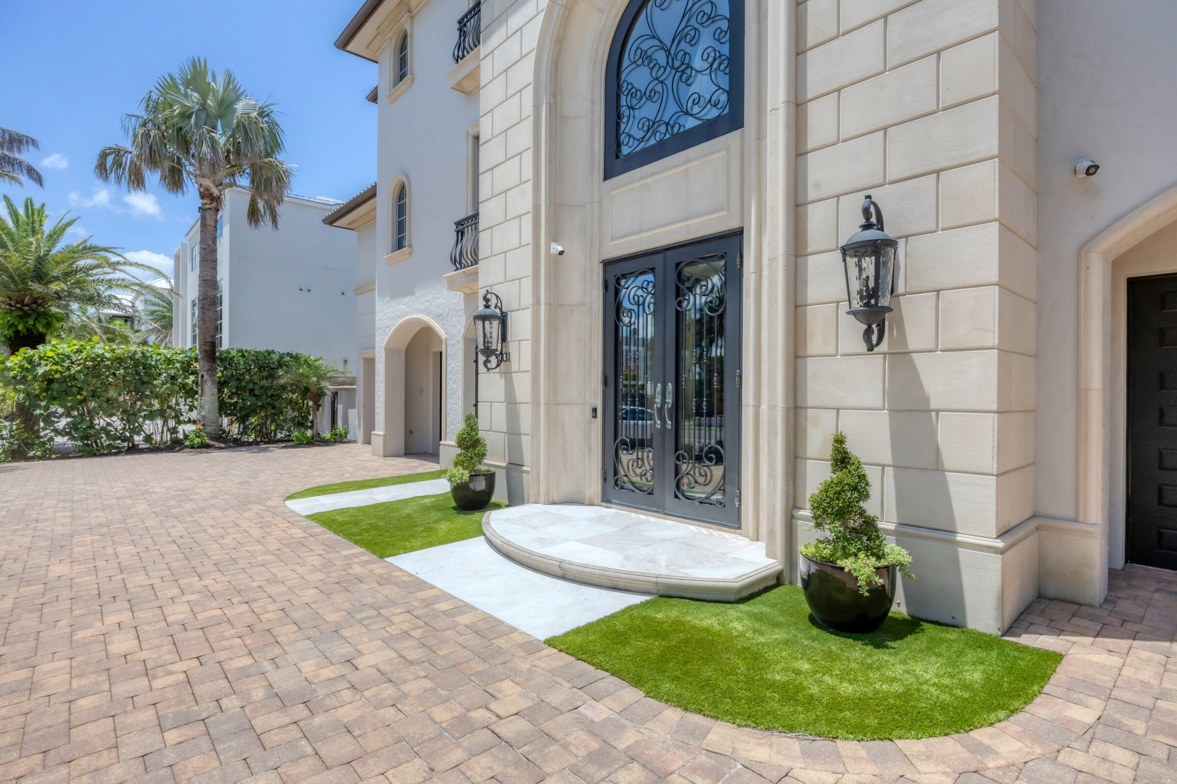 Elegant building entrance with arched doors and wrought-iron detailing, surrounded by manicured landscaping, palm trees, and stone-paved courtyard on a sunny day.
