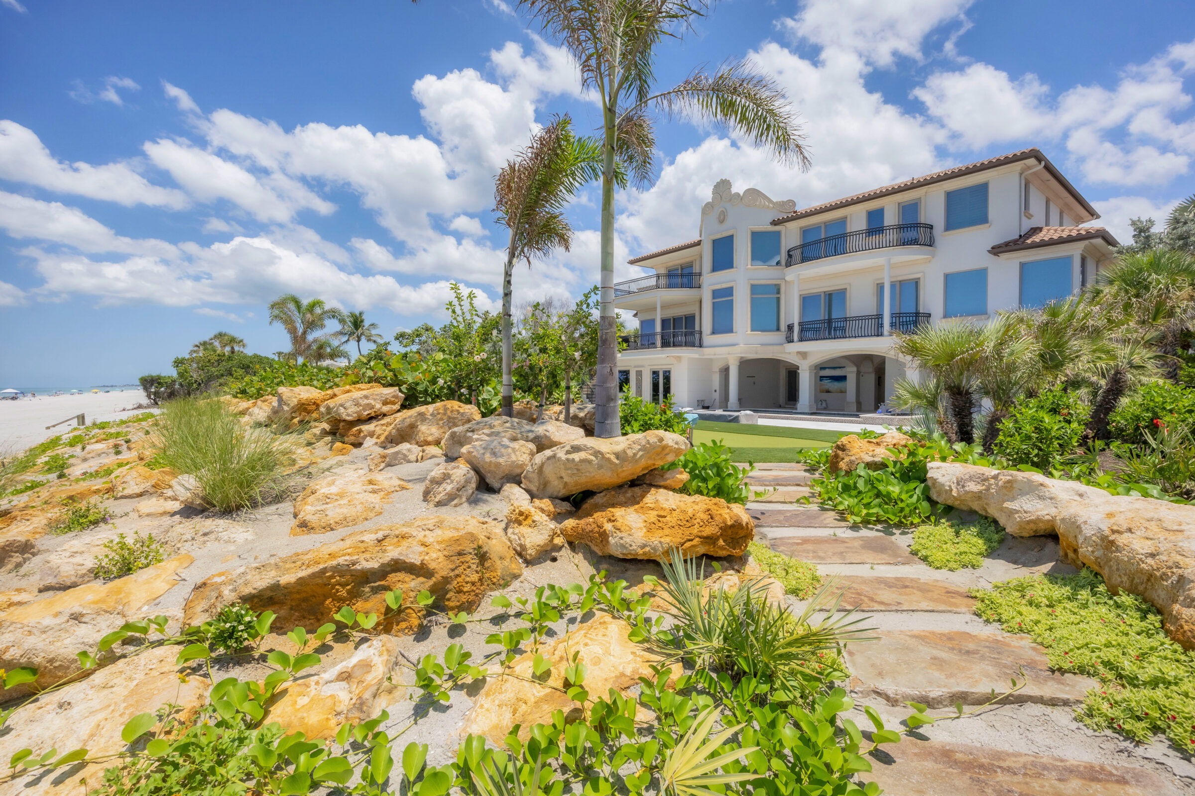 Beachfront property with palm trees, landscaping, and a rocky pathway leading to a large white building, under a clear blue sky with clouds.