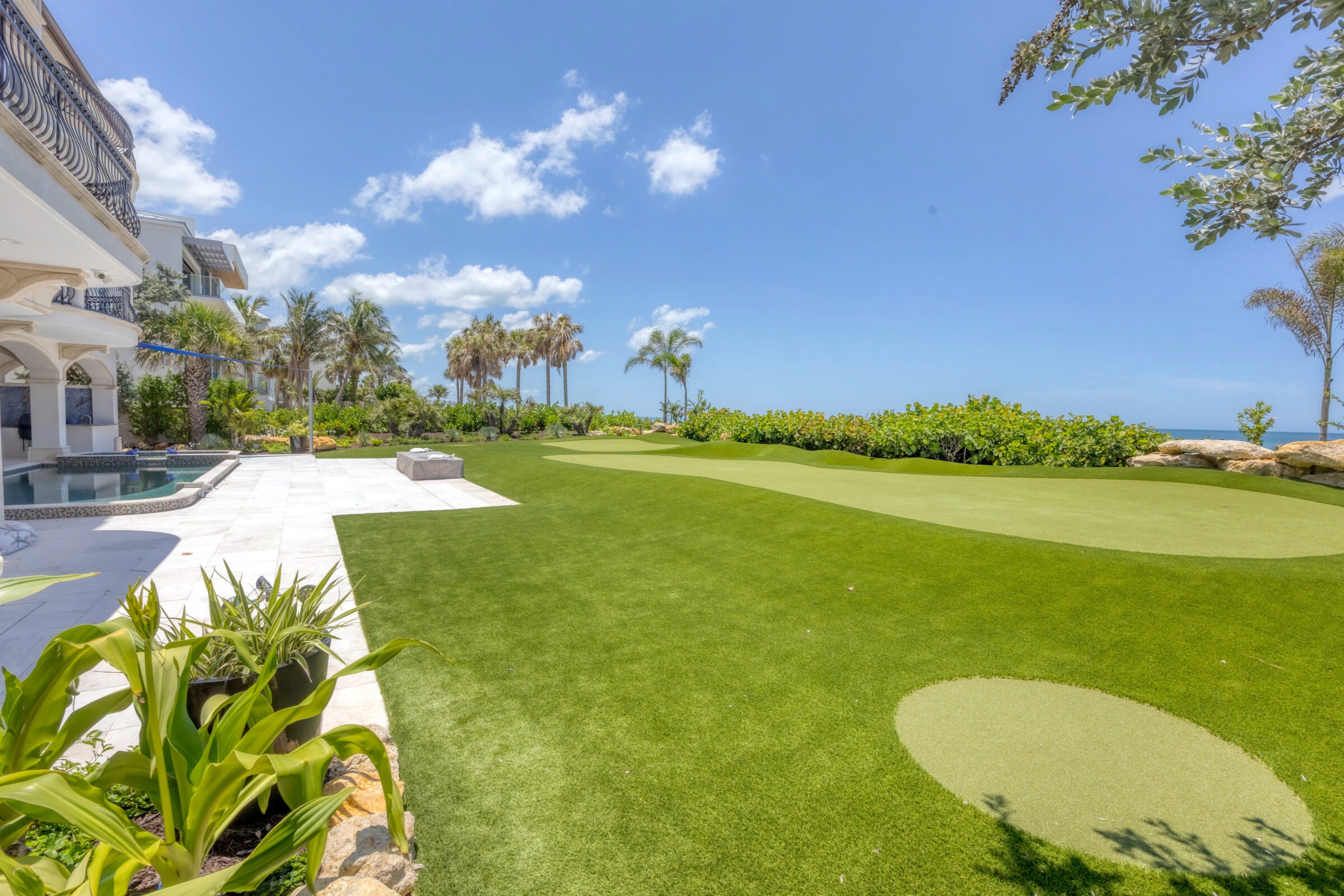 Luxurious villa with a manicured lawn, palm trees, and an elegant pool under a clear blue sky. The ocean is visible in the distance.