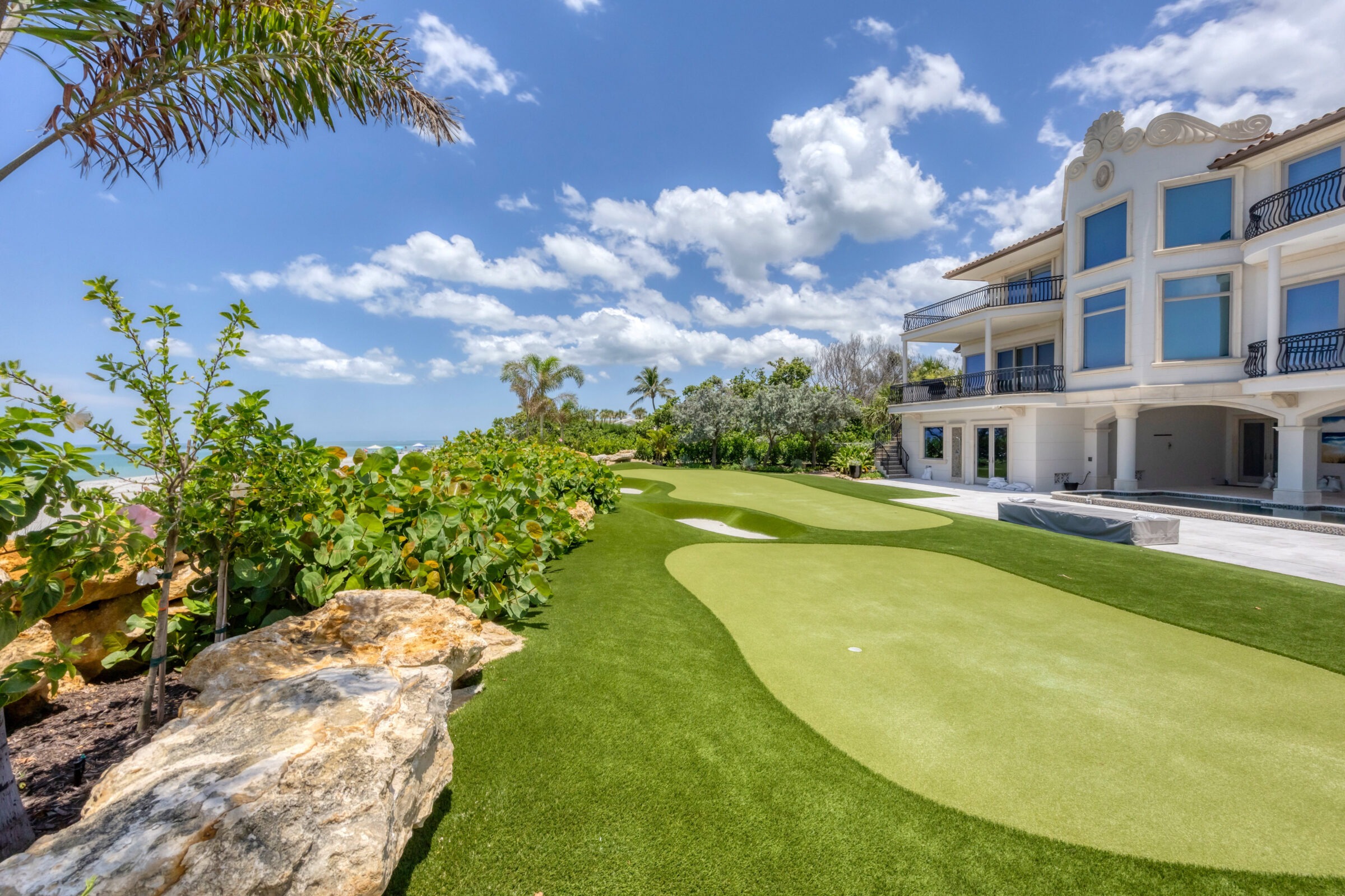 Luxurious beachfront house with manicured lawn, palm trees, and rocky landscaping under a bright blue sky with fluffy clouds by the ocean.