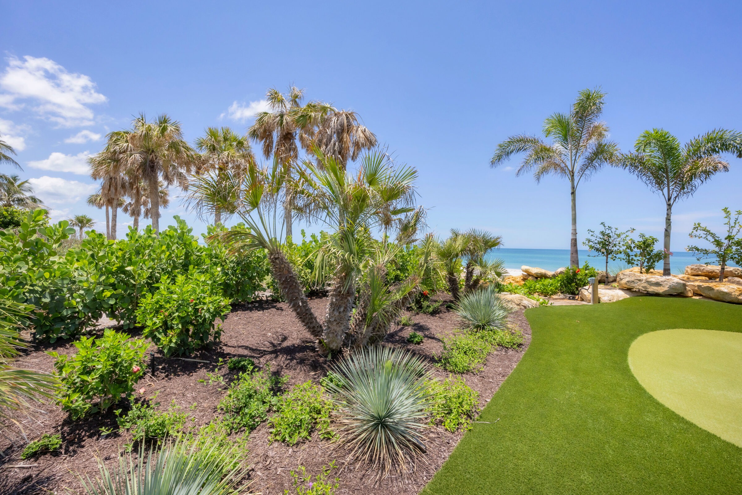 Tropical garden with palm trees, lush greenery, and a small golf putting green overlooking a clear blue ocean under a bright, sunny sky.