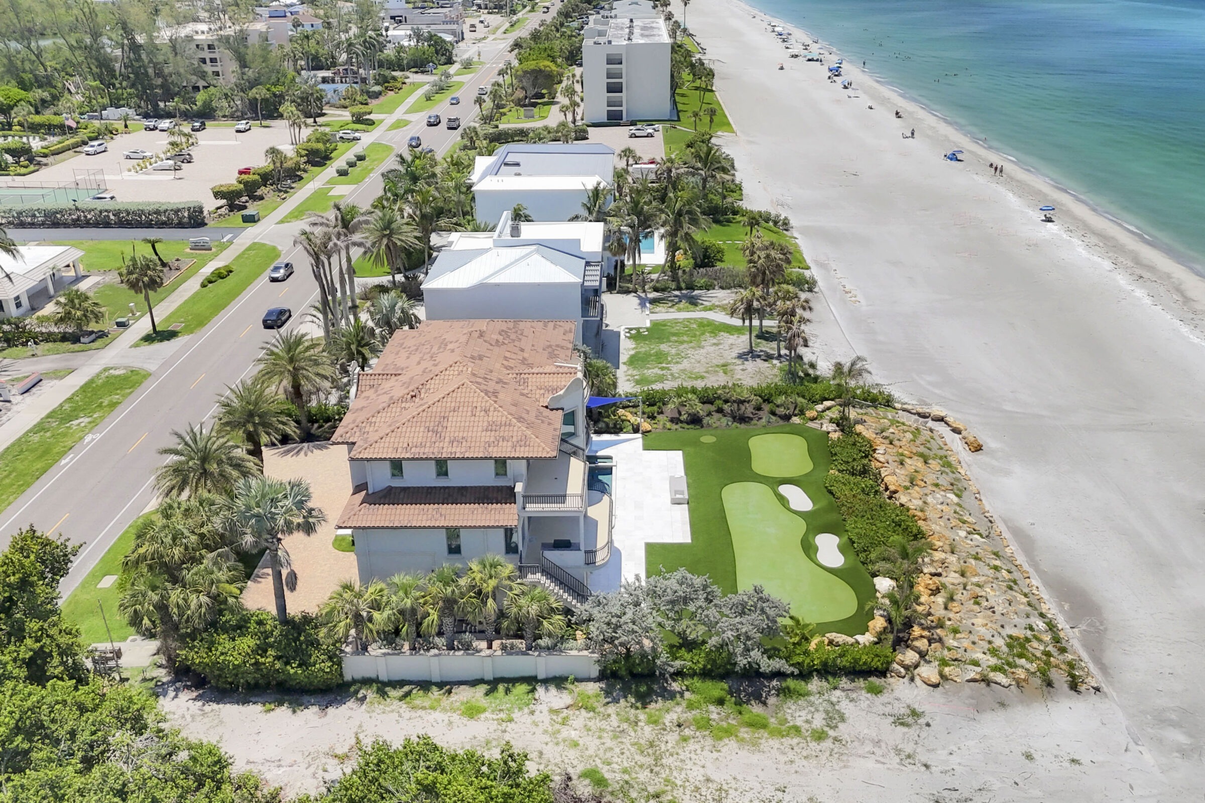 Coastal residential area with modern houses, palm trees, and a white sandy beach. Road parallels shoreline with cars; beachgoers and umbrellas visible.