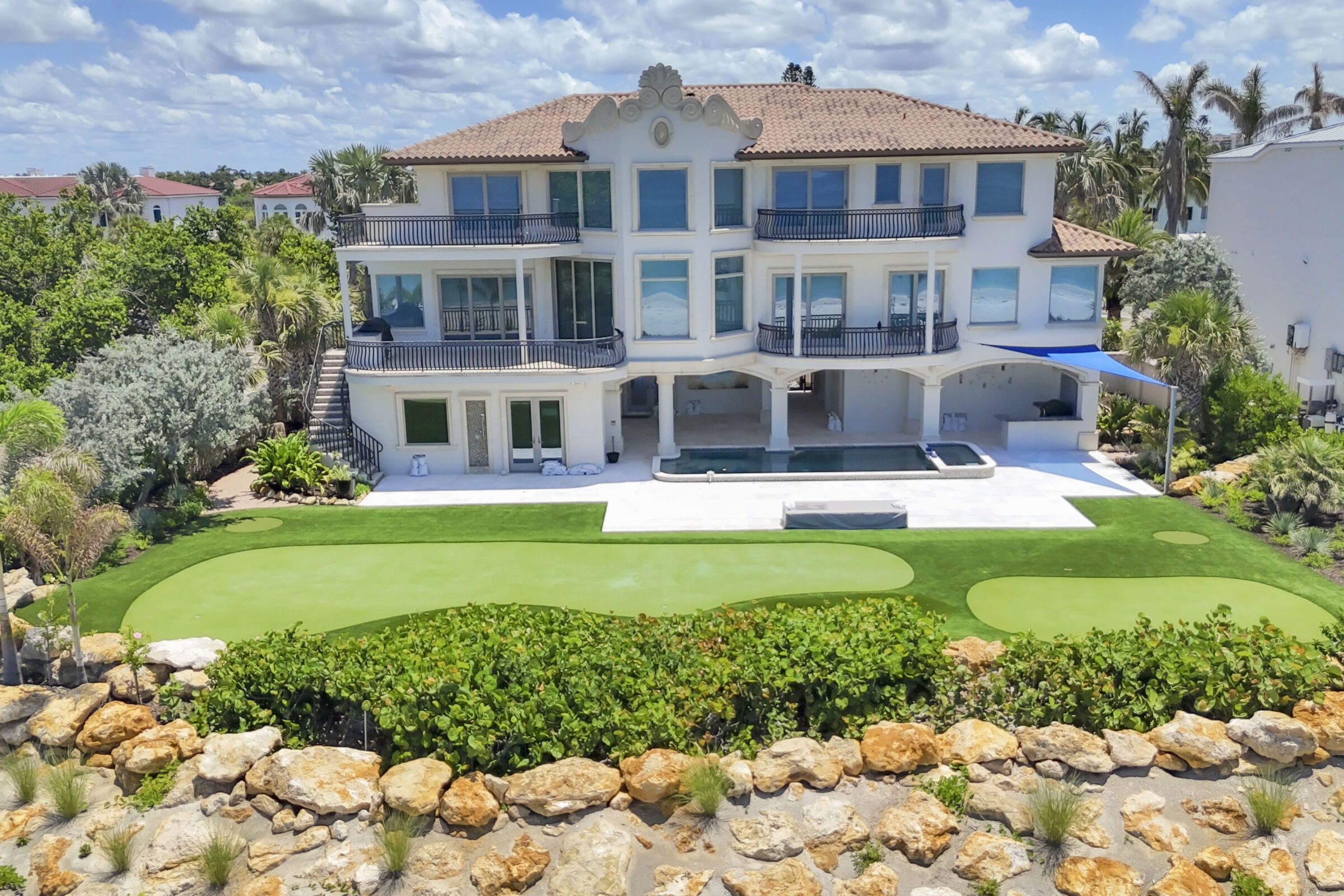 Luxurious three-story villa featuring a terrace, pool, and garden, surrounded by lush greenery and tropical plants under a vibrant blue sky.