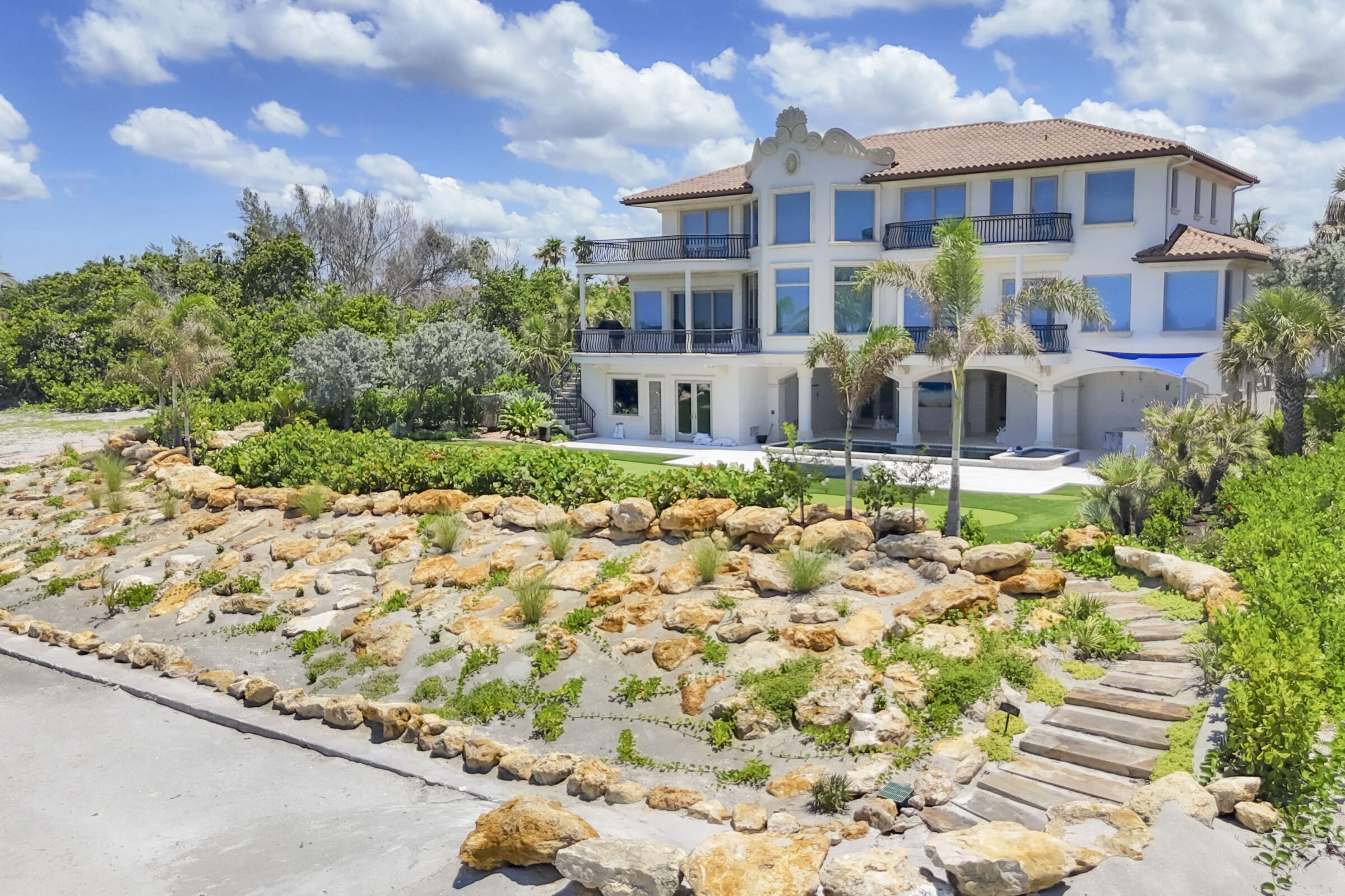 Large Mediterranean-style villa with a terracotta roof, surrounded by lush greenery and stone landscaping, under a vibrant blue sky with scattered clouds.