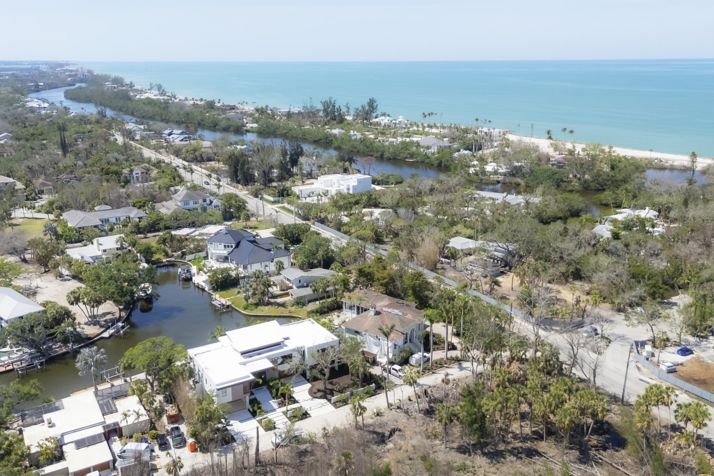 Aerial view of a coastal neighborhood with canals, roads, lush greenery, and sandy beaches along a calm, turquoise ocean under a clear sky.