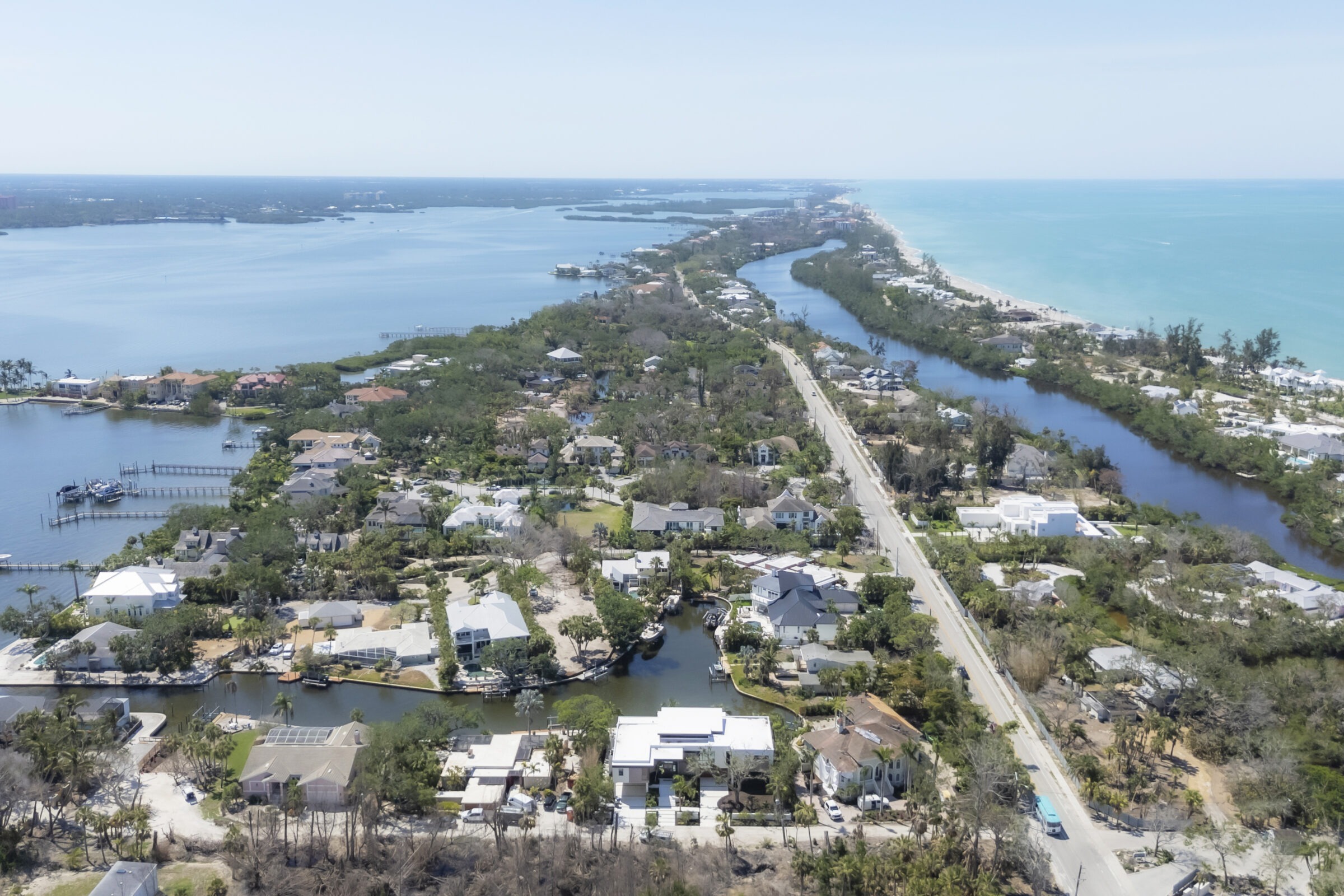 Aerial view of a coastal town featuring waterways, residential houses, dense greenery, and distant ocean views under a clear sky.