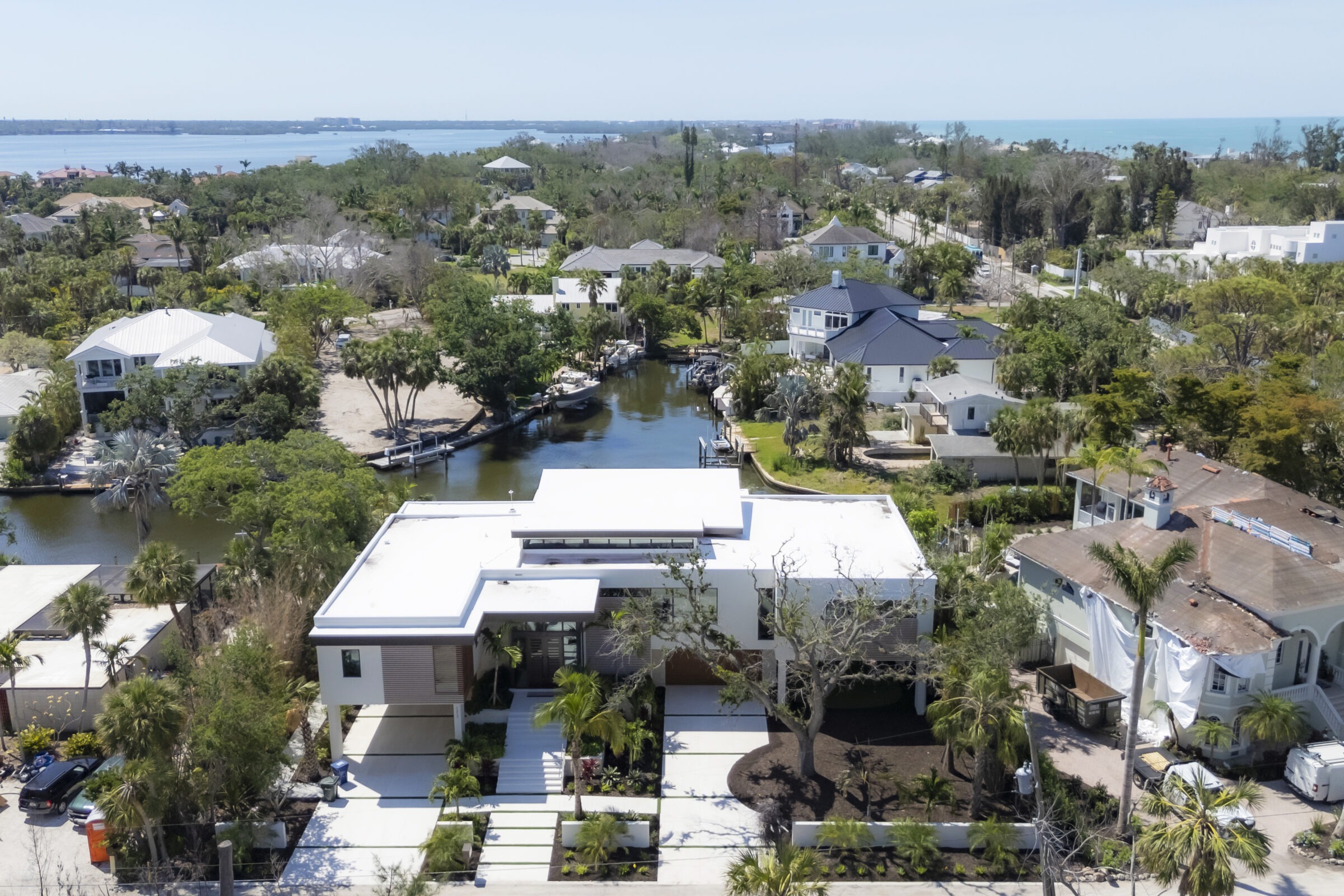 Coastal residential area with modern and traditional houses, surrounded by lush greenery and waterways, leading to a distant ocean view.