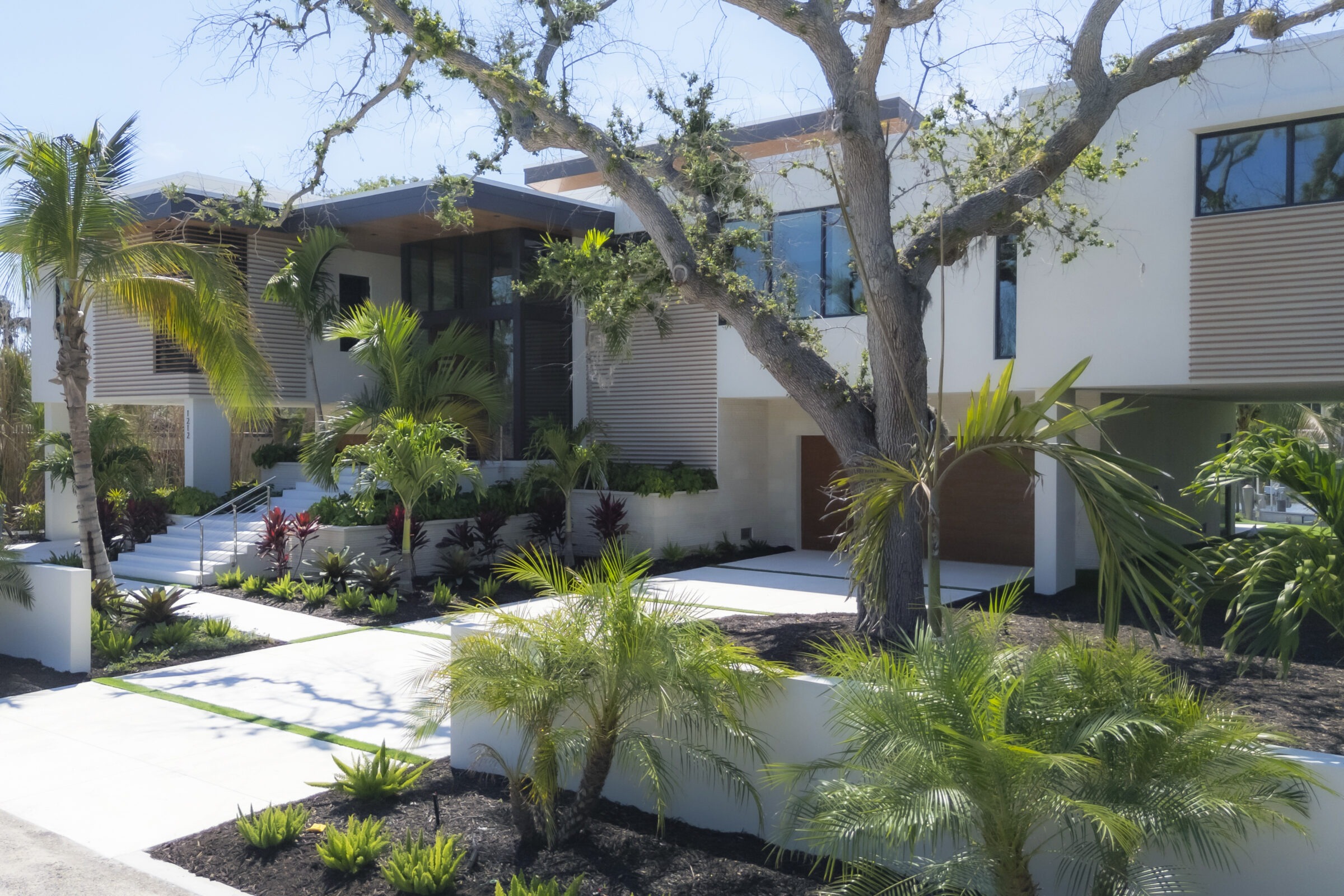 Modern two-story house with a tropical garden, featuring palm trees and shrubs, under blue skies. No recognizable landmarks or historical buildings present.